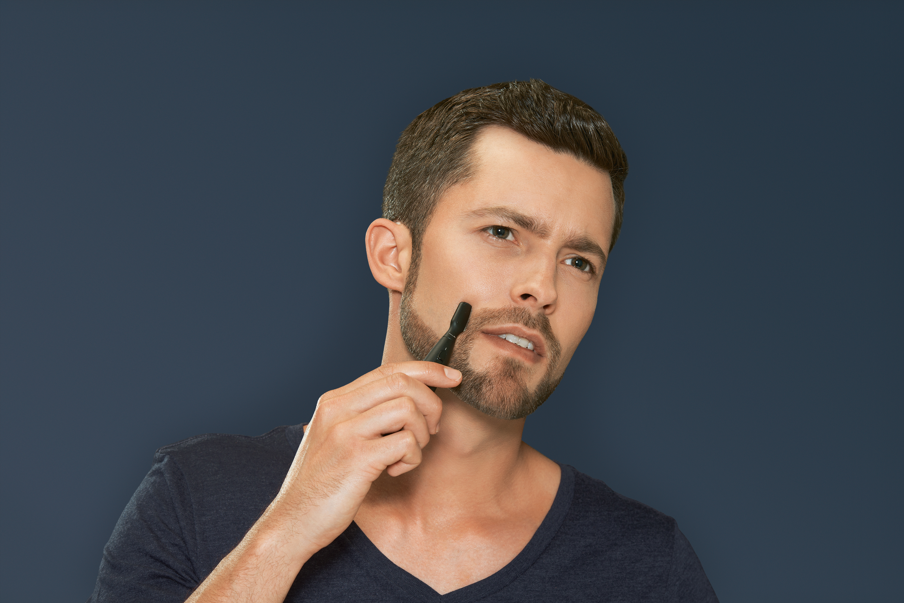 Een man met een baard trimt zijn gezichtshaar met een klein zwart gereedschap, en kijkt nadenkend.