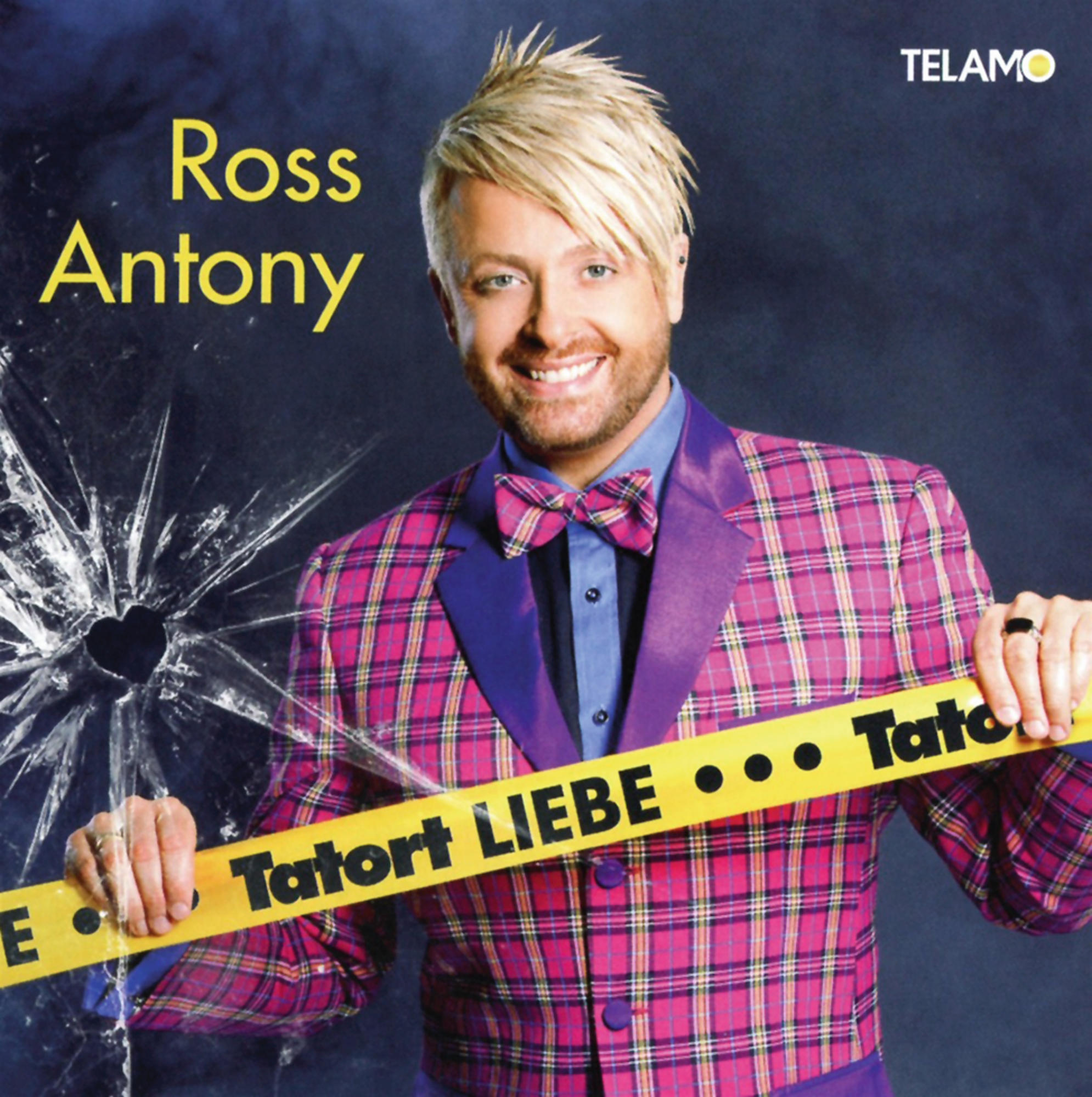 Ross Antony mit blondem Haar, in einer karierten Jacke, hält ein gelbes Klebeband mit "Tatort Liebe".