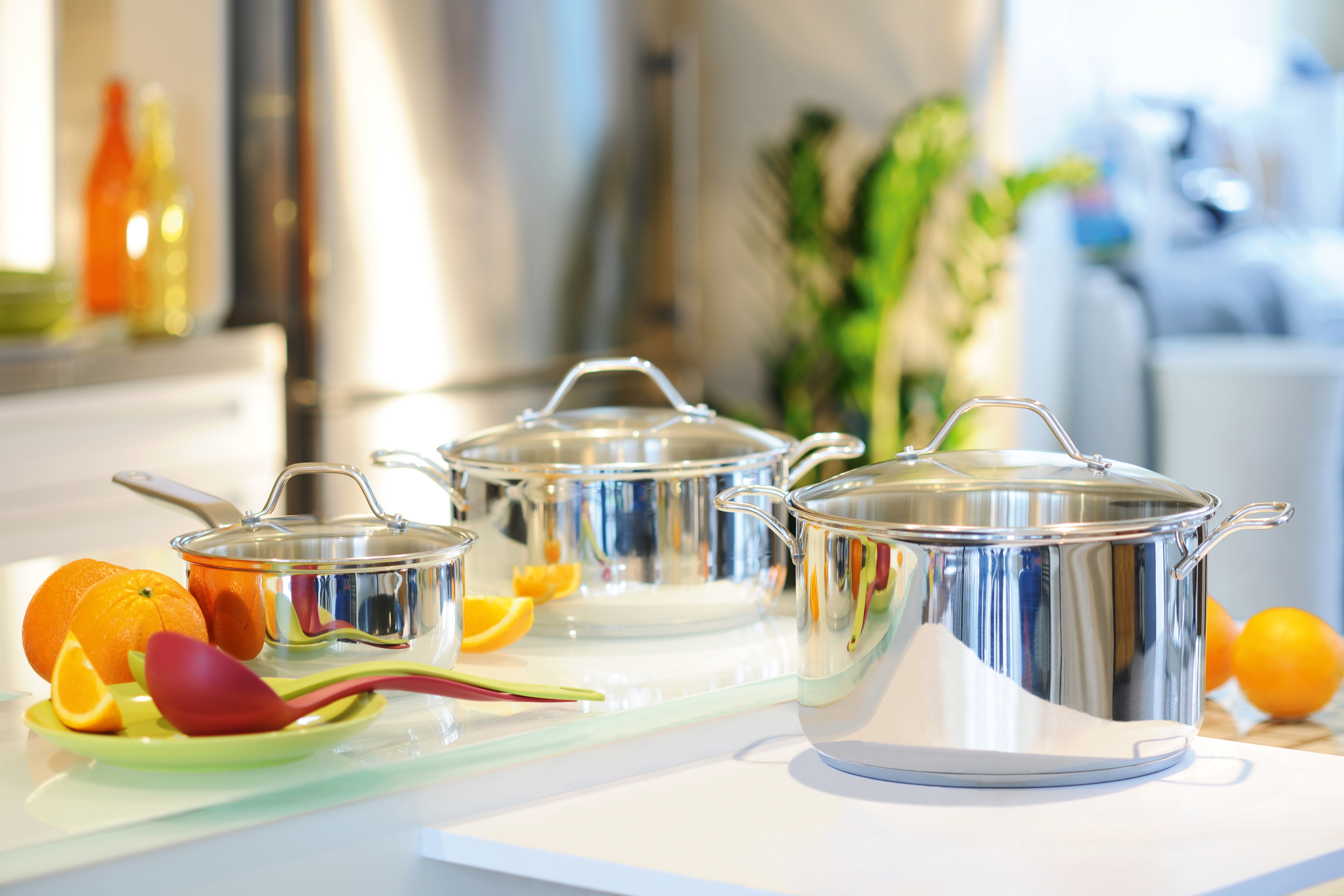 Edelstahltöpfe und -pfannen auf einer weißen Theke. Orangen und ein roter Löffel sind ebenfalls vorhanden.