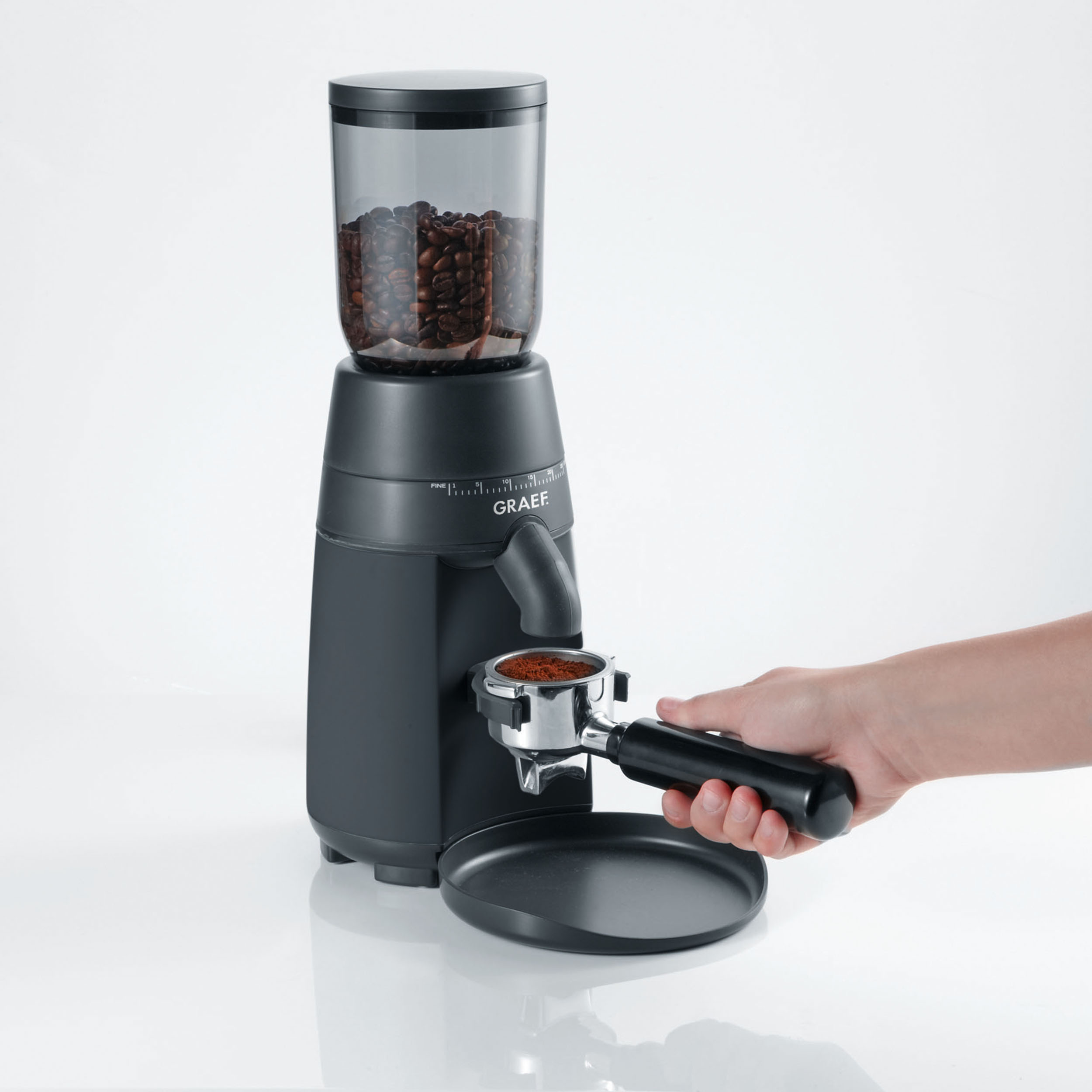 Eine schwarze Kaffeemühle mit Bohnen oben, eine Hand hält einen Siebträger mit Kaffeepulver.