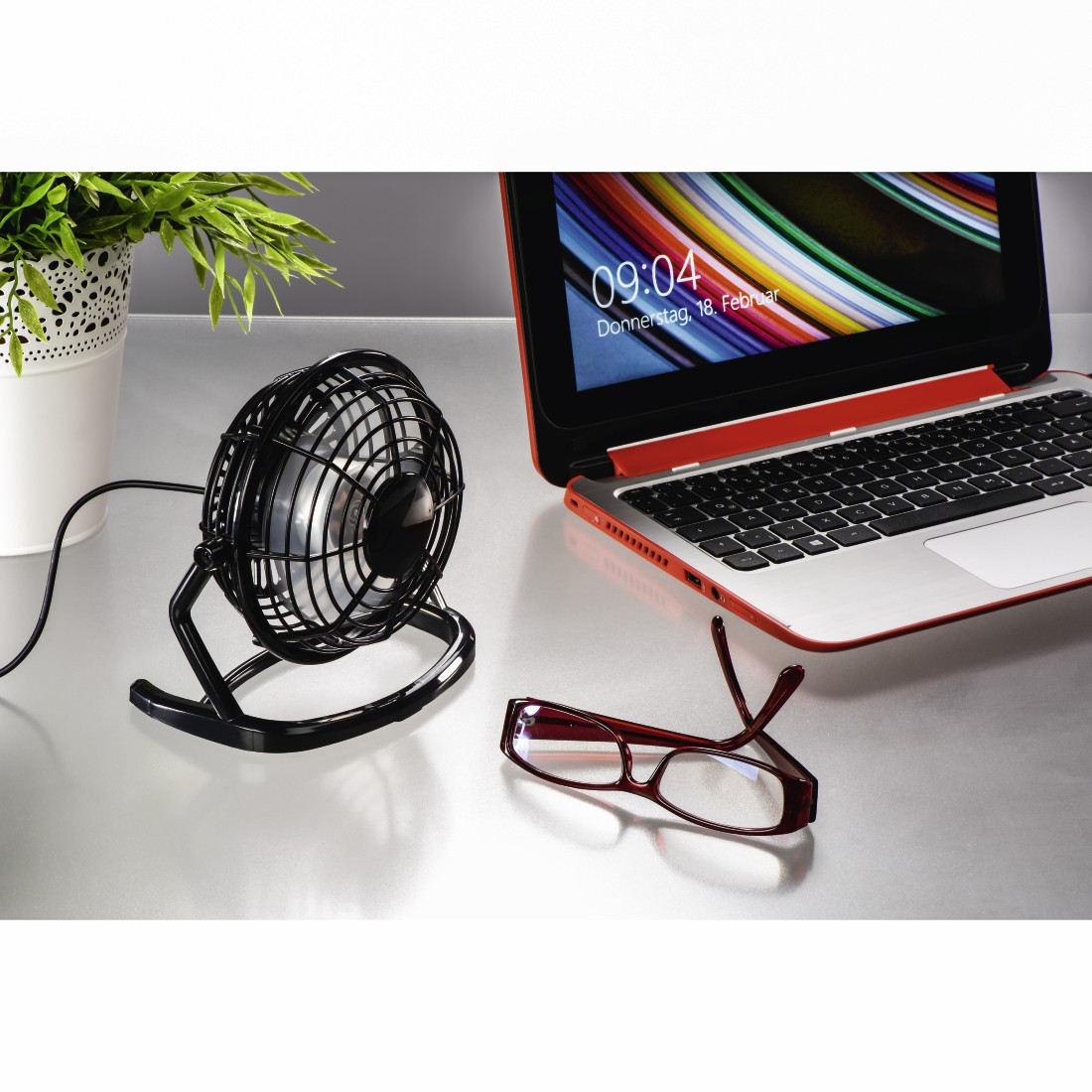 Ein kleiner Tischventilator, ein Laptop und eine Brille auf einer silbernen Oberfläche.