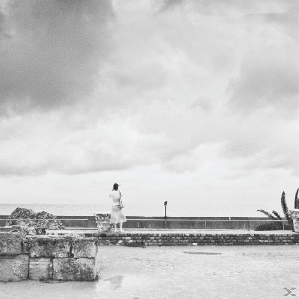 Eine Person steht auf einer Steinmauer und blickt unter einem bewölkten Himmel auf das Meer.