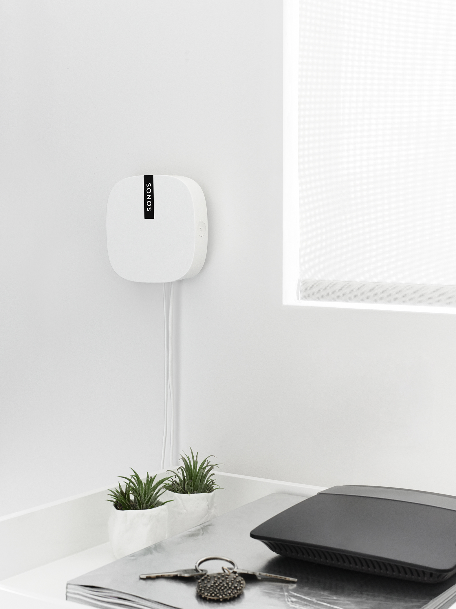 Appareil Sonos blanc sur mur blanc, avec deux petites plantes et un routeur.