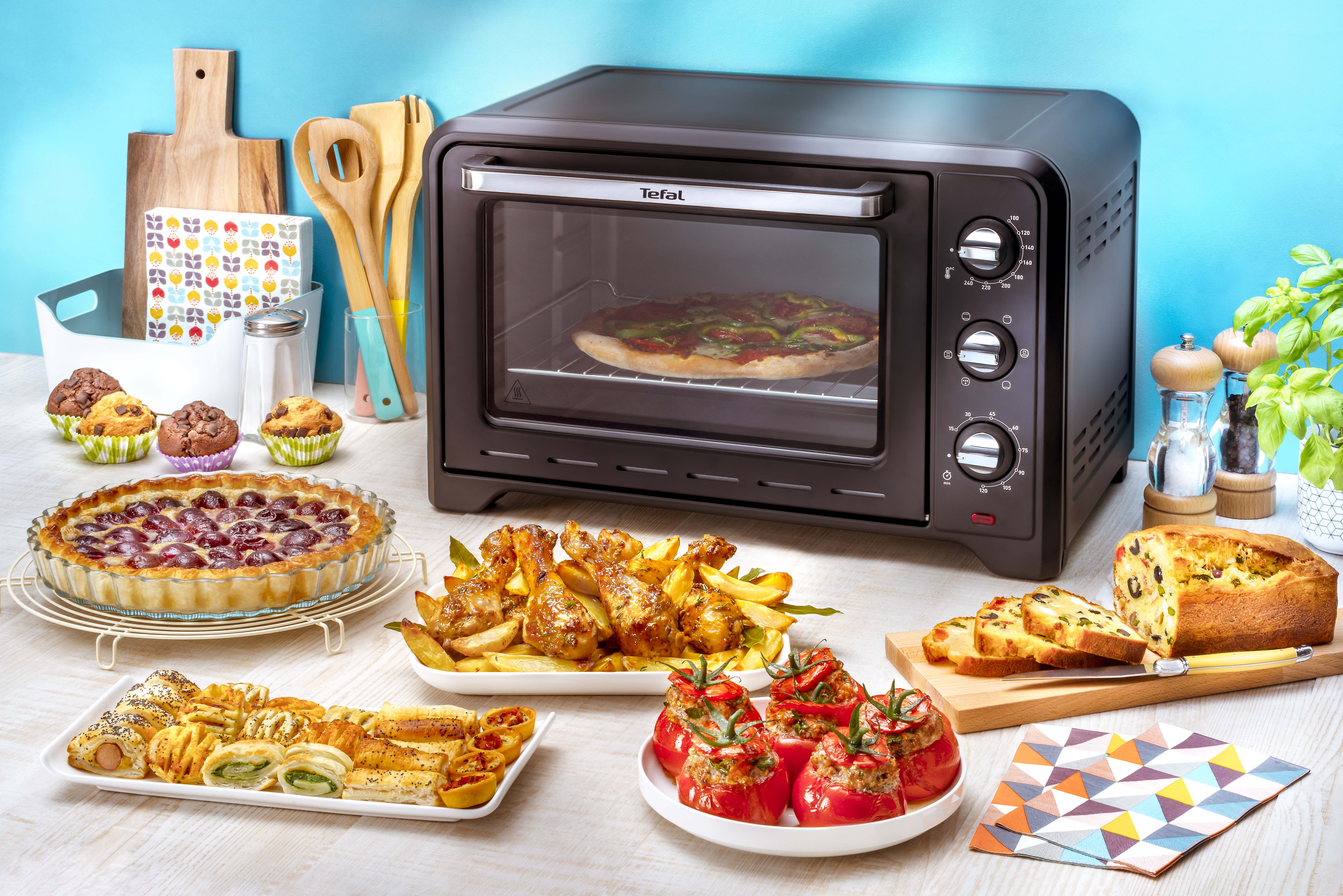 Een oven met pizza erin, omringd door eten. Cakes, taarten en gebraden voedsel op een tafel.