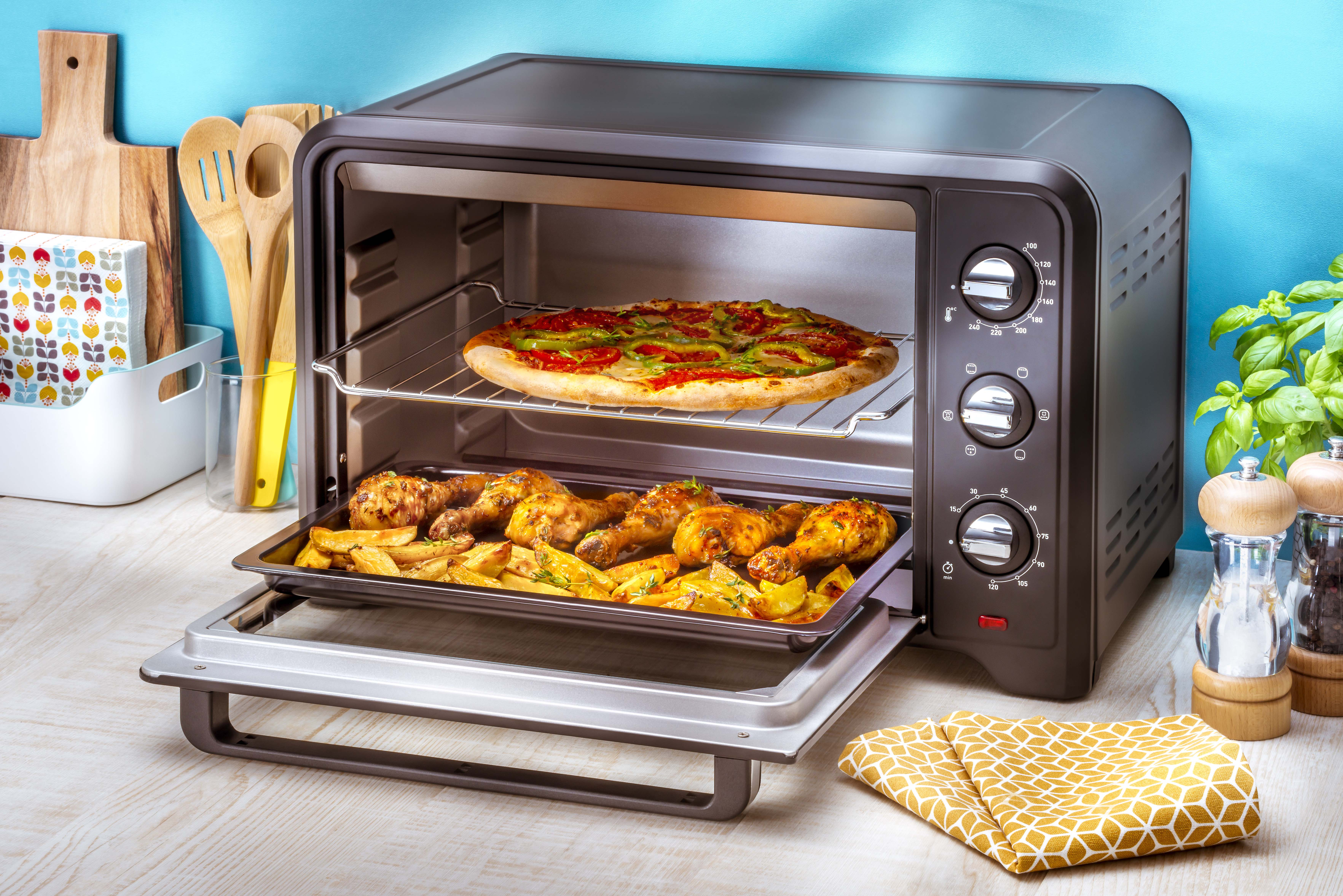 Een open zwarte oven met pizza en kip. Een gele theedoek en houten keukengerei liggen in de buurt.