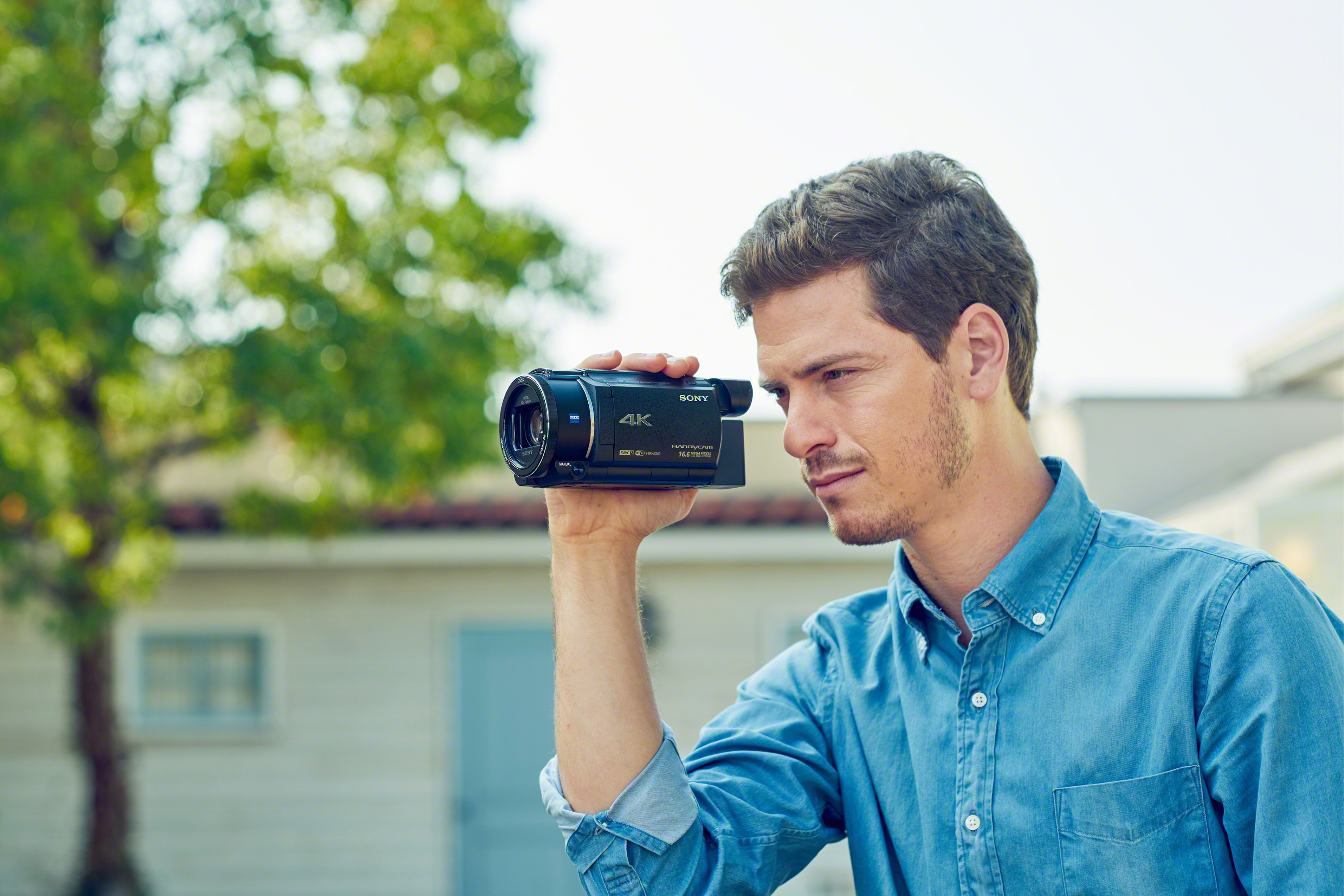 Un uomo tiene una videocamera nera, guardando lo schermo. Un albero verde è sullo sfondo sfocato.
