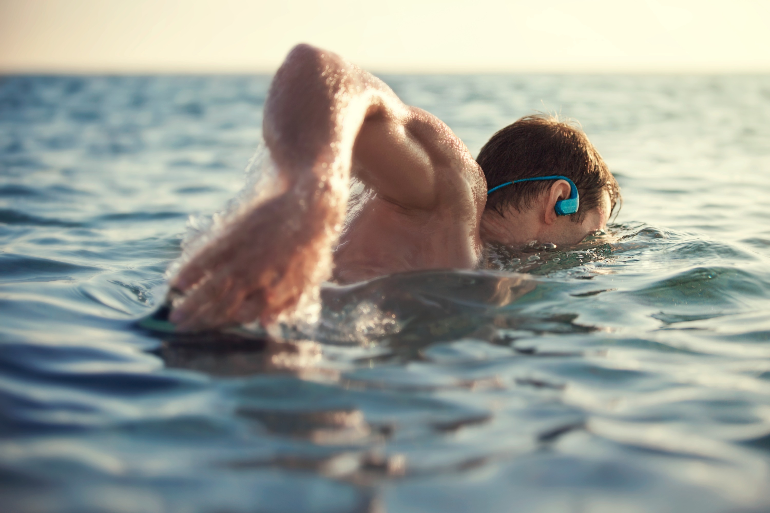 Mann schwimmt in blauem Wasser. Er trägt einen blauen Kopfhörer. Das Wasser ist ruhig und spiegelt den Himmel.