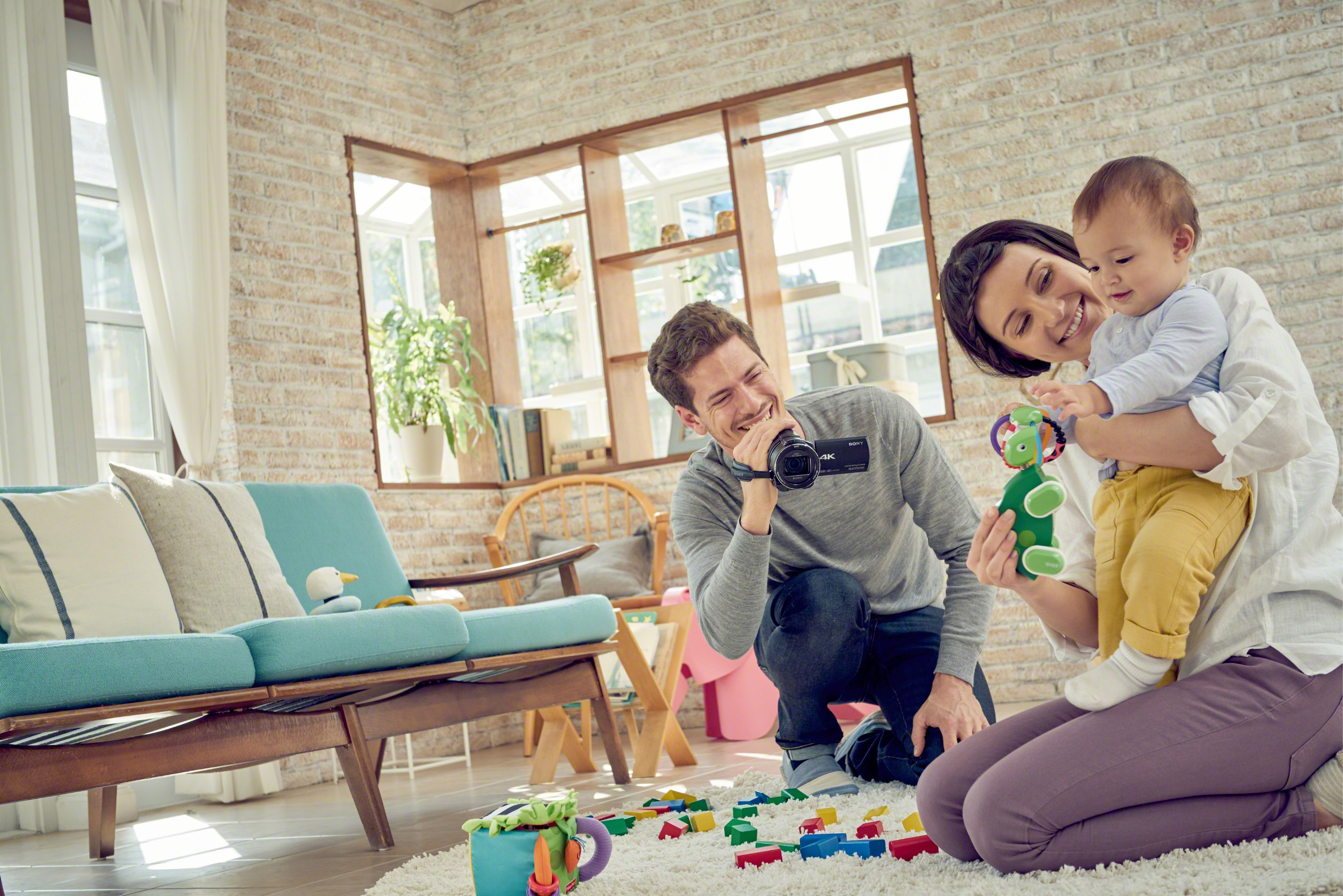 Famiglia in soggiorno, padre che filma con una telecamera, madre che tiene in braccio il bambino. Giocattoli sparsi.