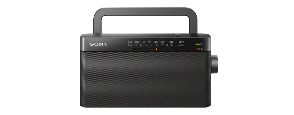Siyah bir Sony radyo, tutma kolu var. Hoparlör ızgarası, ayar kadranı ve frekans göstergesi var. Arka plan beyaz.