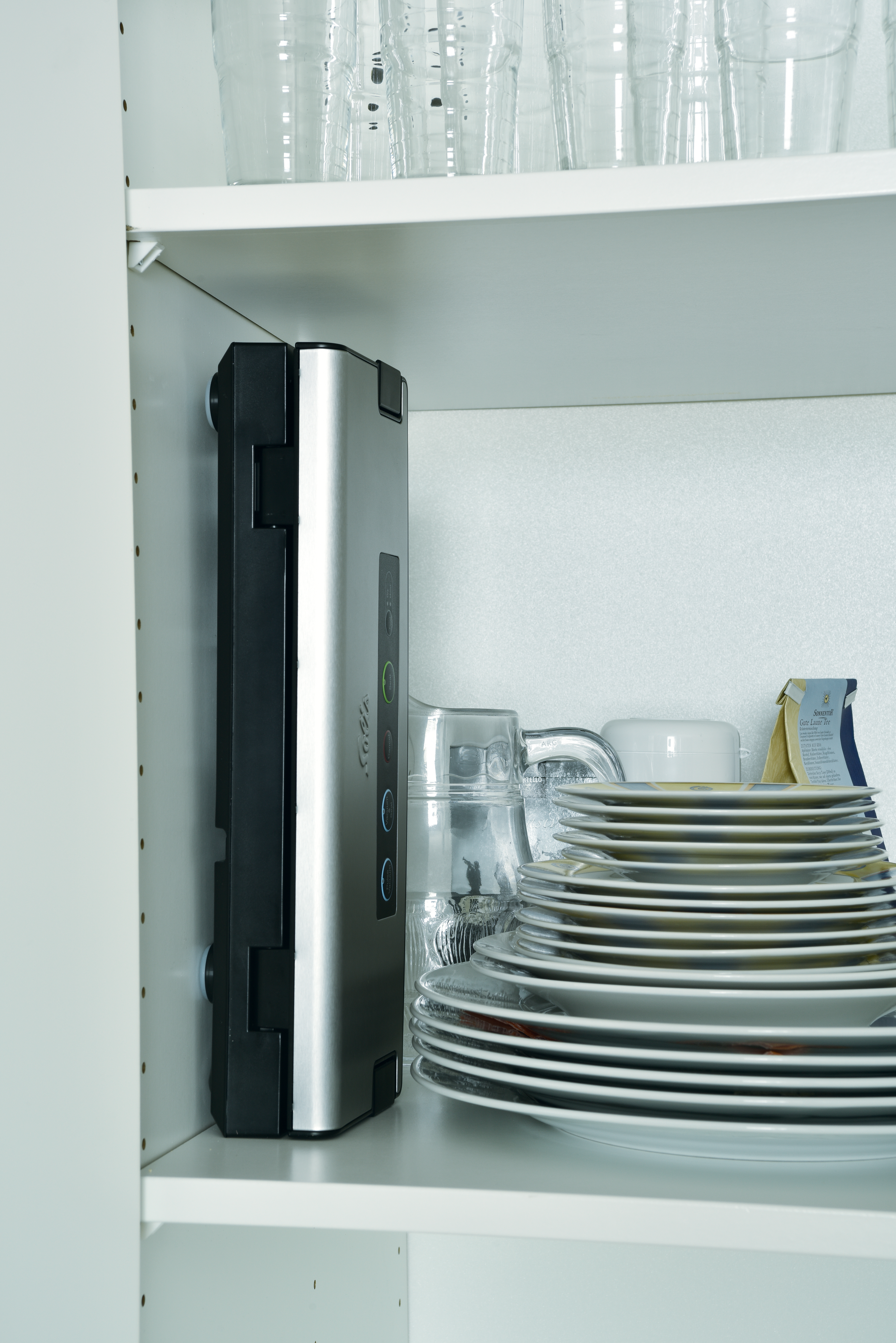 A kitchen cabinet shelf with a silver vacuum sealer, glassware, plates, and a small container. White background.