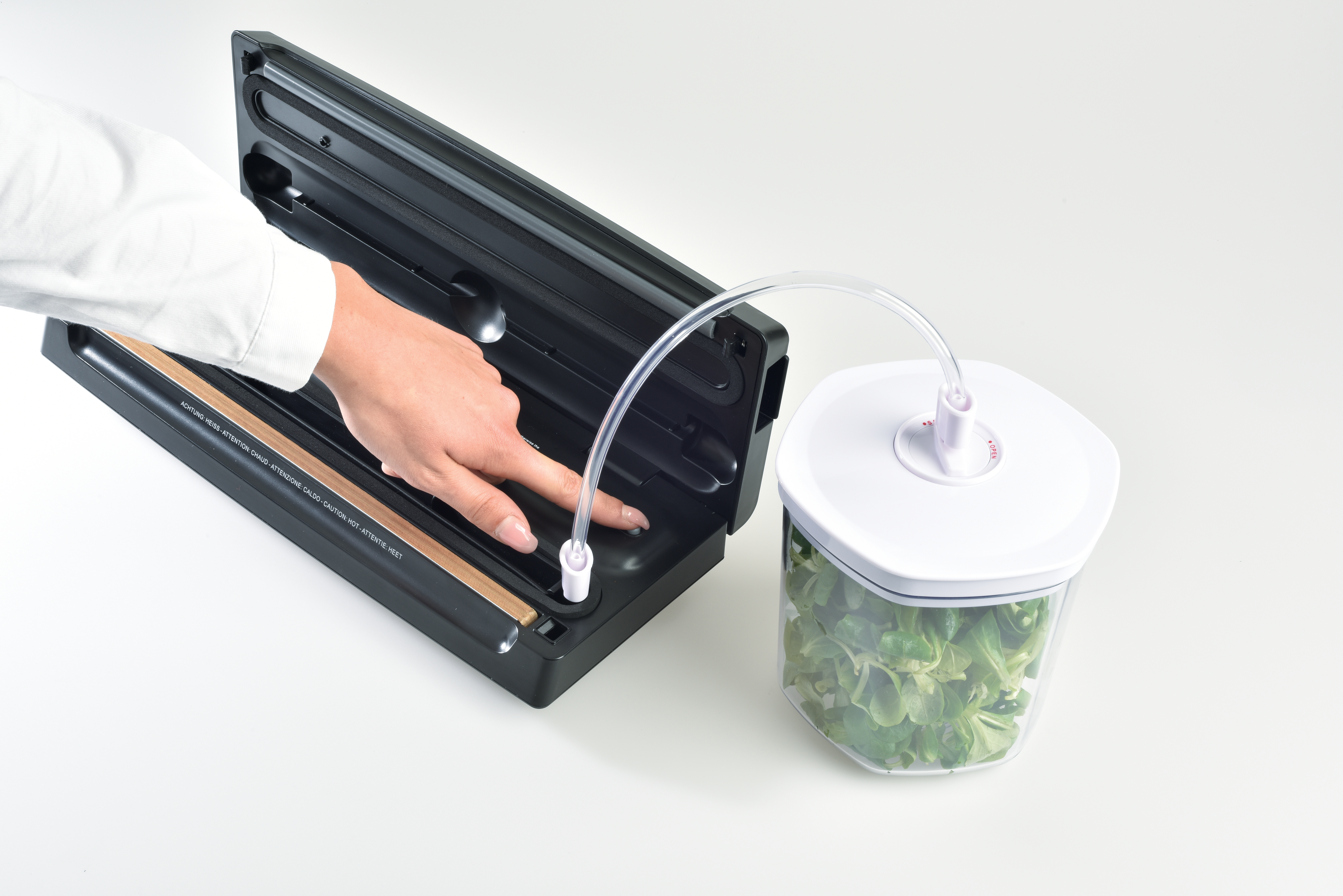 A hand operates a black vacuum sealer connected by a tube to a container filled with green leaves, on a white surface.
