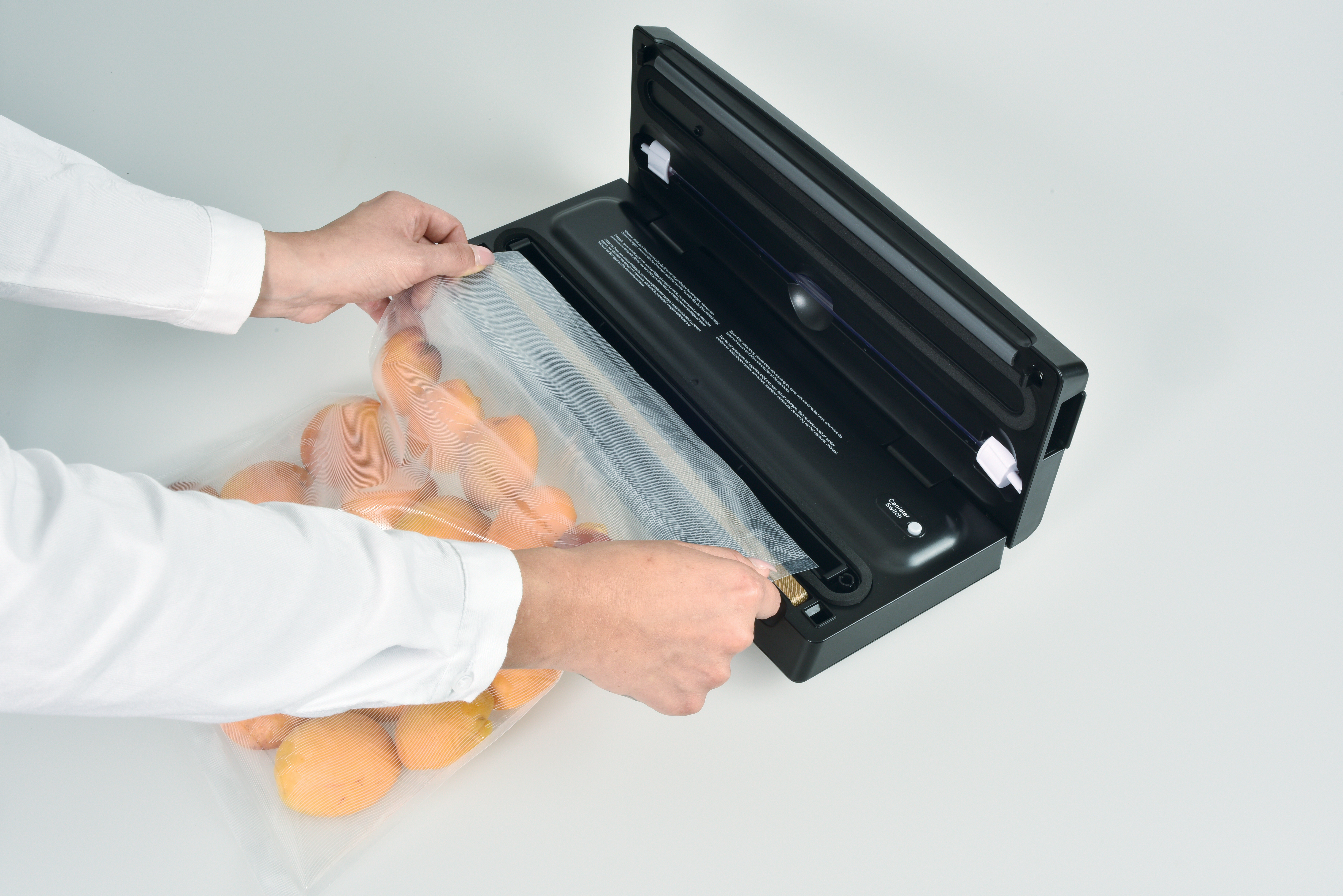 Hands hold a bag of produce, placing it into a black vacuum sealer. Oranges are in the bag. White surface background.