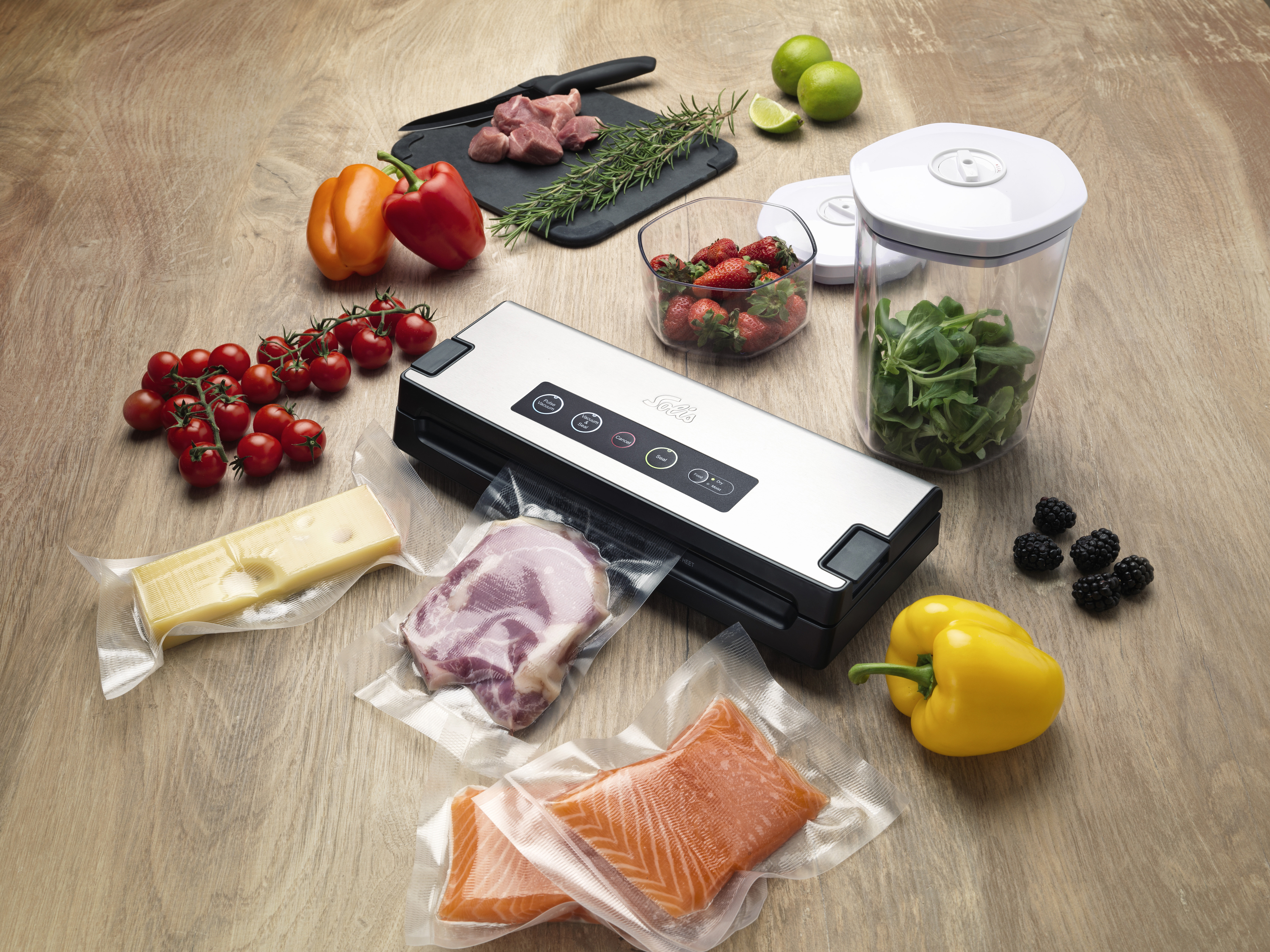 A wooden table with a vacuum sealer, various foods like meat, fish, vegetables, and fruits, and a black cutting board.