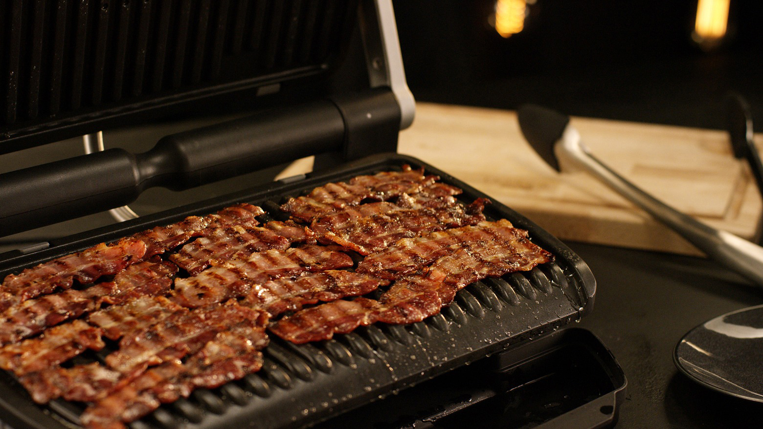 Speck brät auf einem schwarzen Grill. Ein Pfannenwender und ein Teller sind daneben.