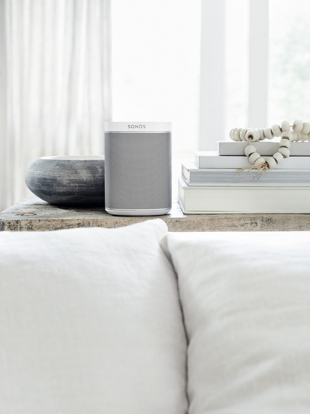 Ein weißer Sonos-Lautsprecher, Bücher und eine Vase stehen auf einem Holztisch. Weiße Kissen sind im Vordergrund.