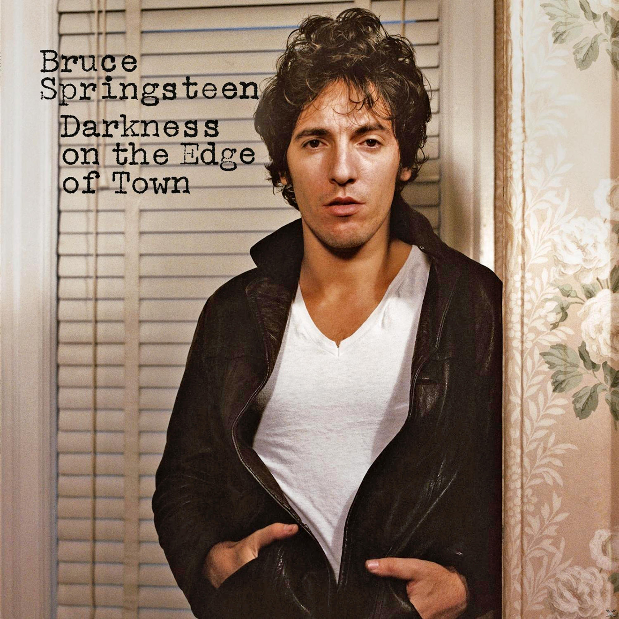 Bruce Springsteen steht. Er trägt eine schwarze Jacke über einem weißen Hemd. Der Hintergrund ist eine Tür.