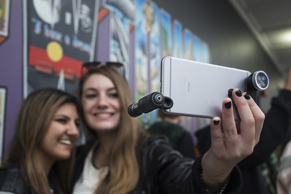 Zwei lächelnde Frauen machen ein Selfie mit einem Telefon und Aufsteckobjektiv vor einem bunten Wandbild.