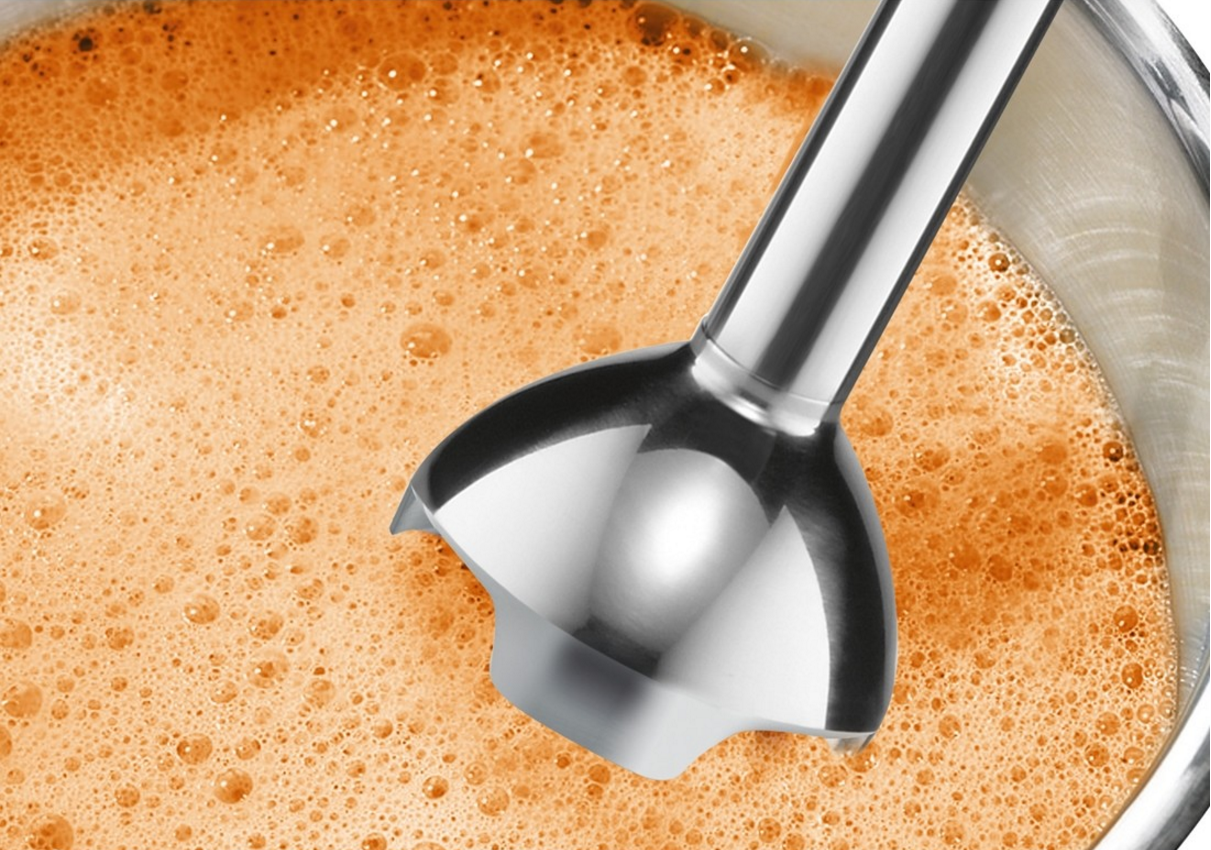 Gros plan d'un mixeur plongeant argenté dans un liquide orange, à l'intérieur d'un bol.