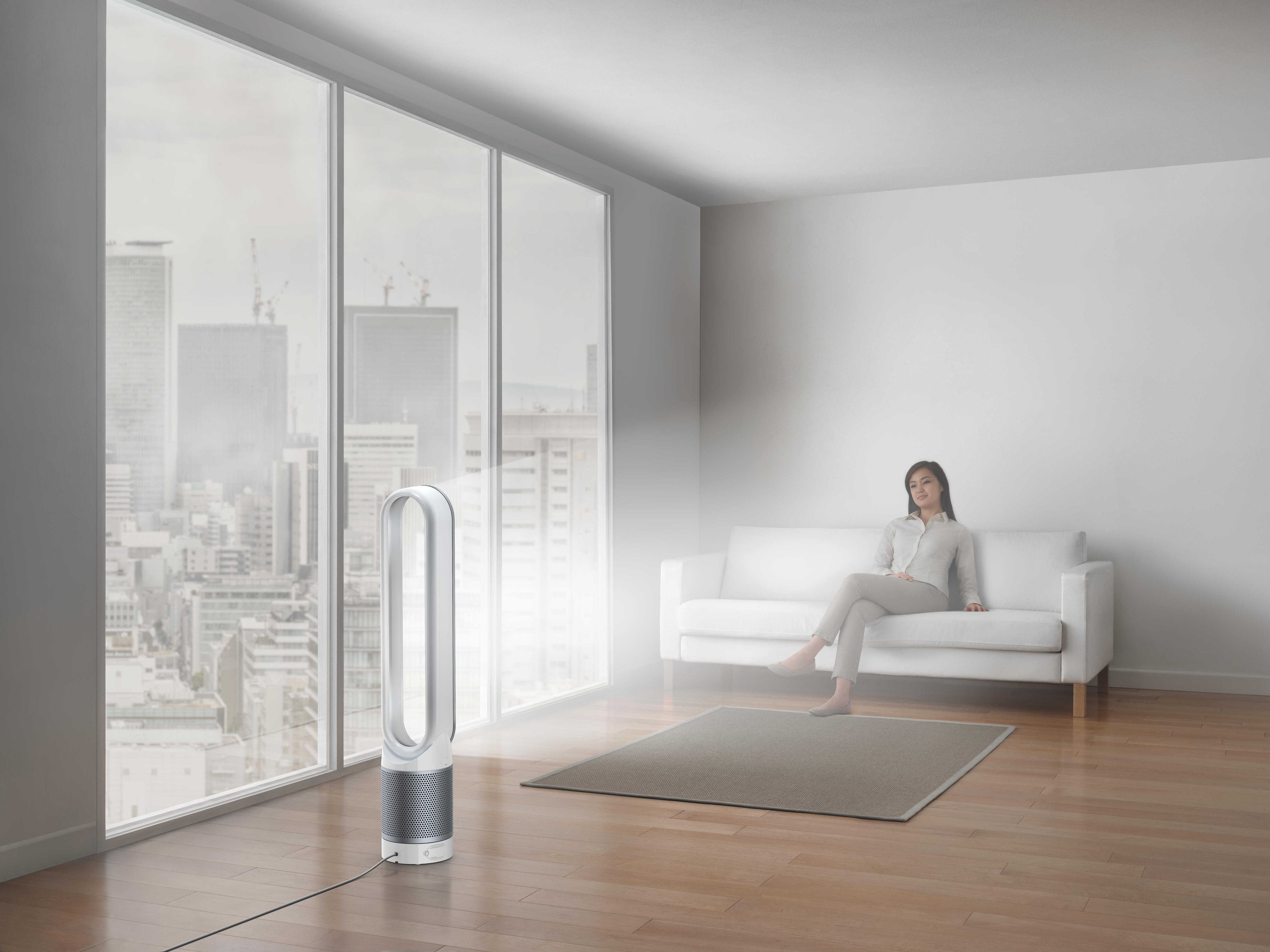 Eine Frau sitzt auf einem weißen Sofa in einem modernen Raum. Ein Ventilator steht auf dem Boden.