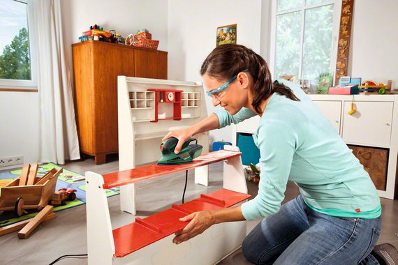 Nő egy szobában, polcot szerel elektromos szerszámmal, védőszemüveget visel. Bútorok és játékok a háttérben.