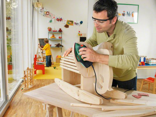 Egy férfi fa hintalovat csiszol elektromos csiszolóval, bent egy gyerekkel.