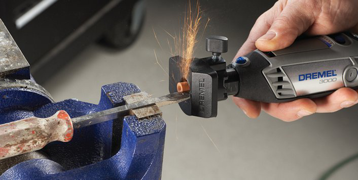 Egy kéz Dremel szerszámot használ egy kék satuba fogott fémtárgyon, szikrák repülnek.