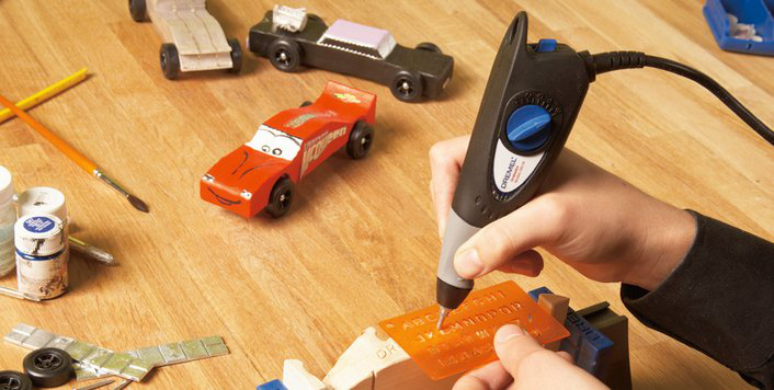 Kezek Dremel szerszámot használnak betűk gravírozásához egy faautóra, más játékautók a közelben.