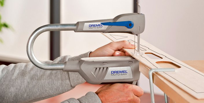 Egy személy Dremel Moto-Saw-val vág egy darab fát. A fűrész szürke és kék.