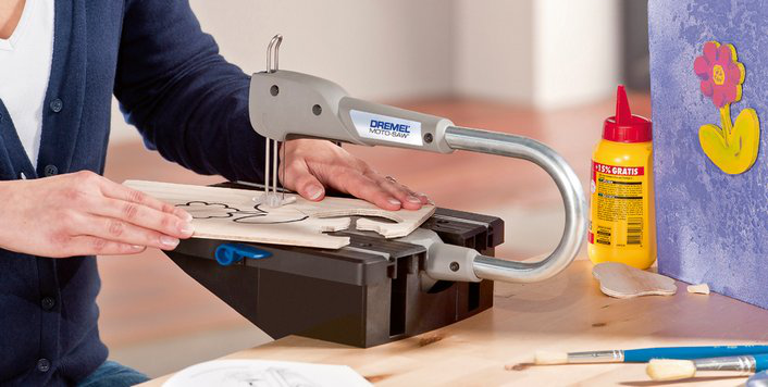 Egy személy Dremel Moto-Saw-t használ egy faforma kivágásához egy munkapadon, ragasztóval és szerszámokkal a közelben.