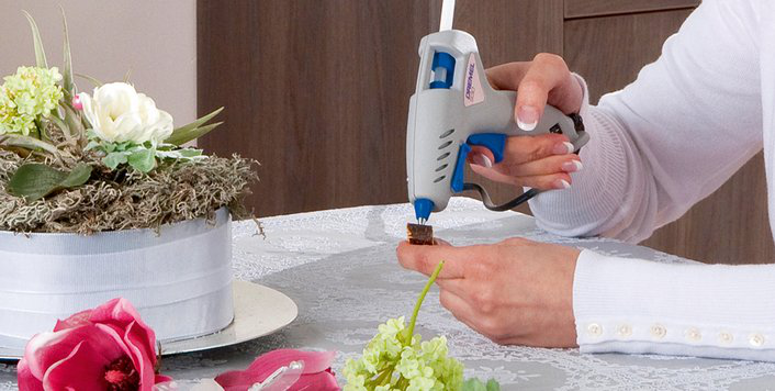 Egy személy Dremel ragasztópisztollyal ragaszt egy tárgyat. Virágok, fehér asztal.