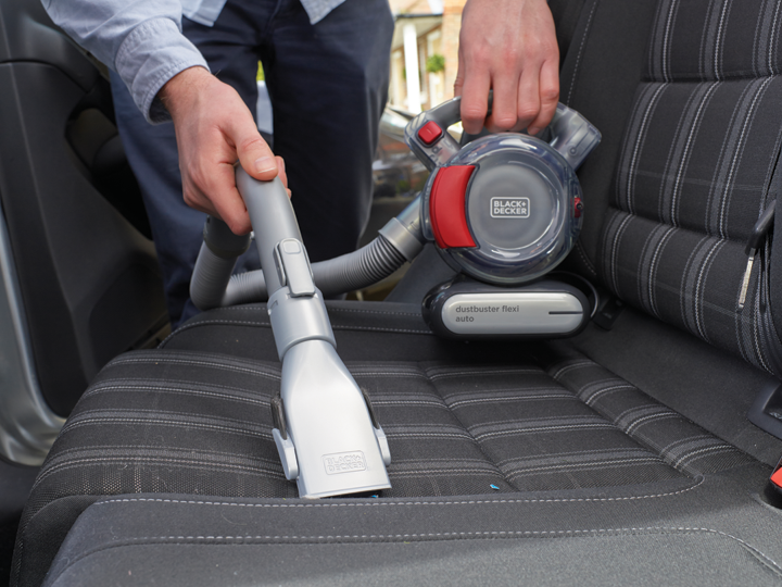 Egy kéz egy Black+Decker porszívót tart, amely egy fekete autóülést tisztít. A porszívó piros és fekete.