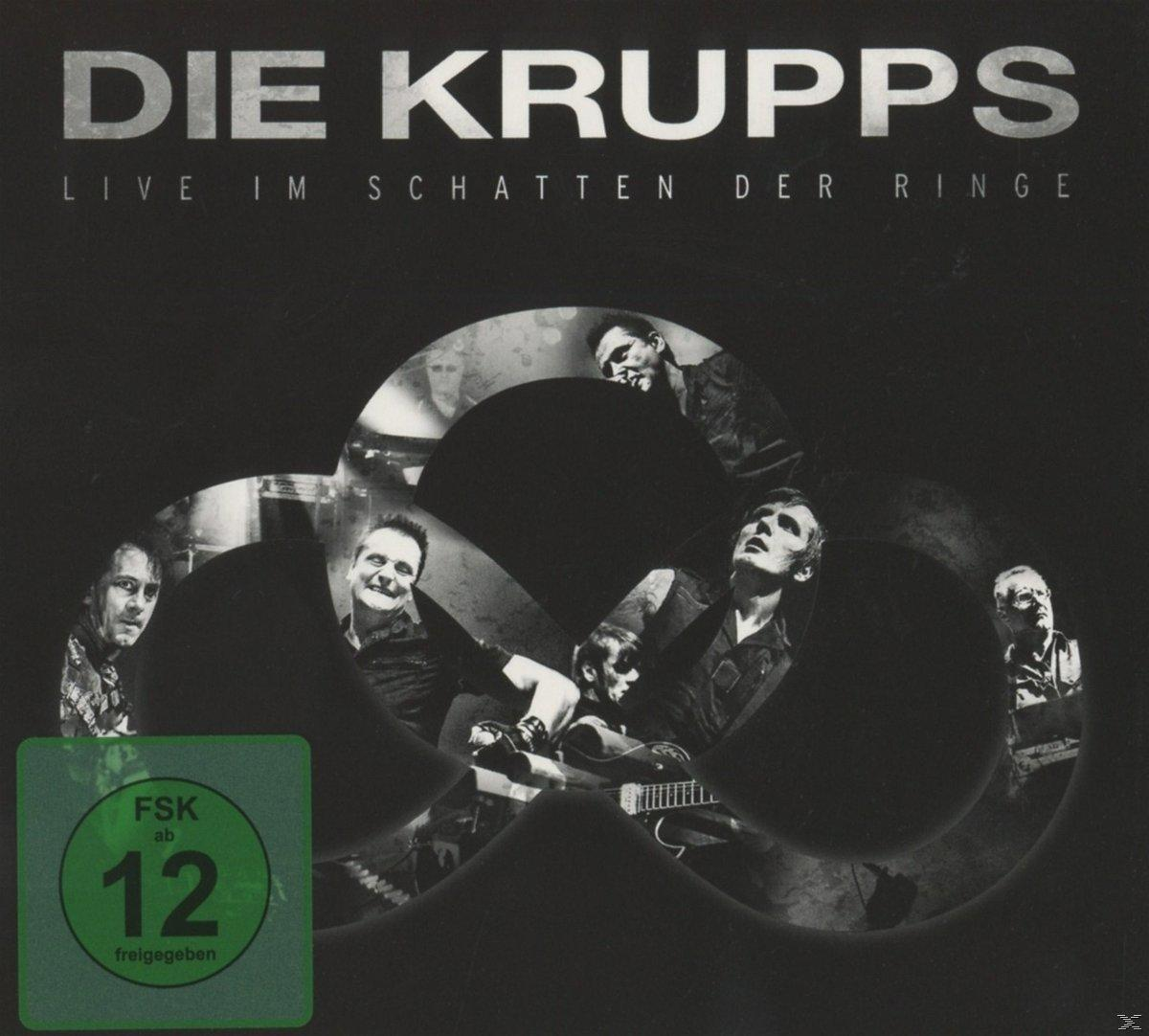 CD-Cover: Die Krupps, Live im Schatten der Ringe. Die Band ist in dunkler Umgebung mit grünem Label zu sehen.