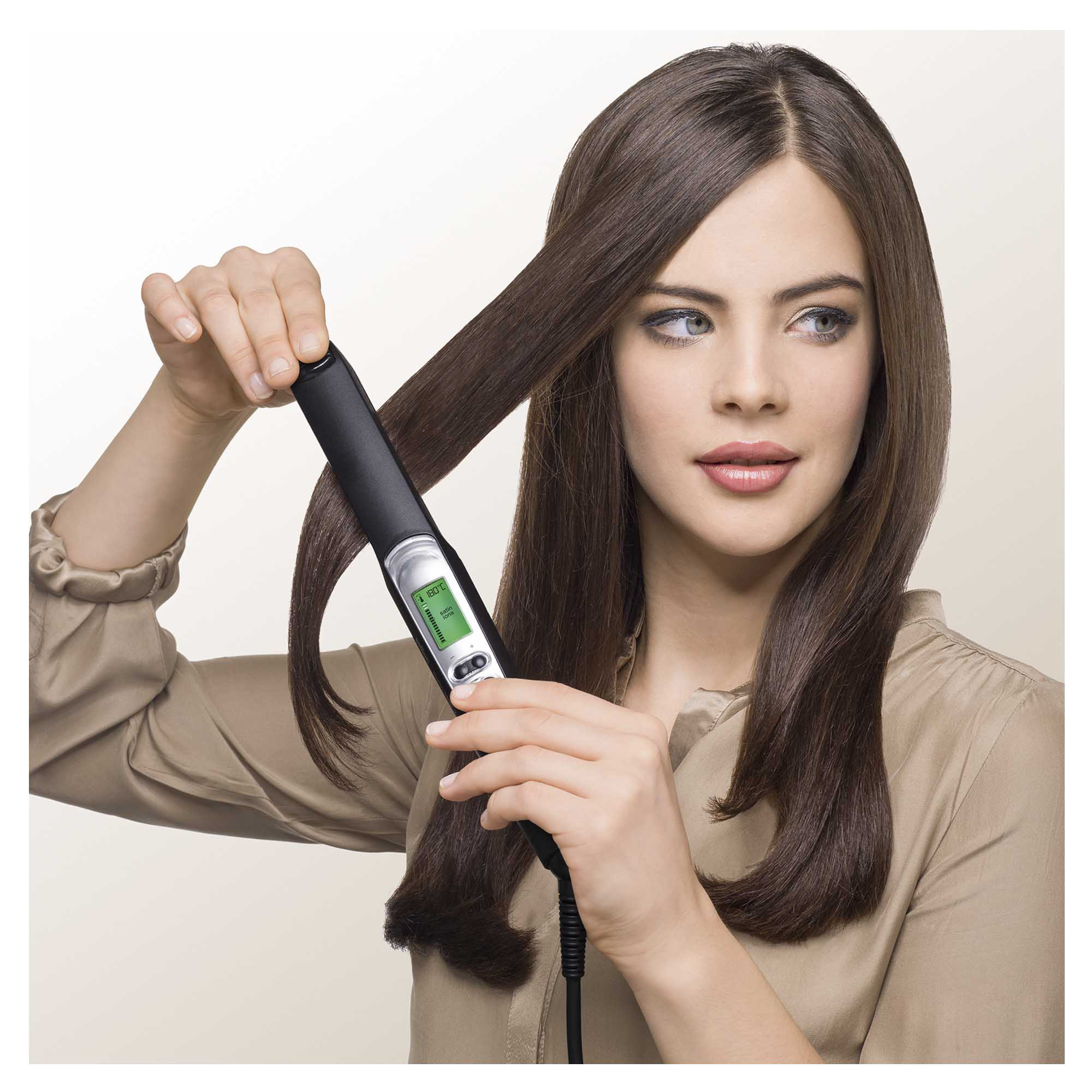 Eine Frau benutzt einen schwarzen Haarglätter auf ihrem braunen Haar. Sie hält eine Haarsträhne und blickt weg.