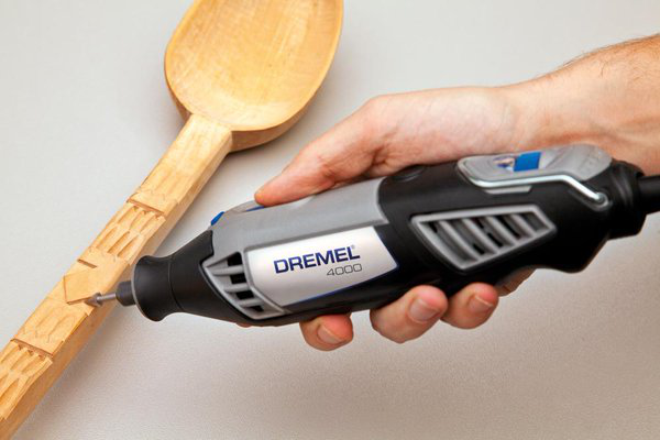 Egy személy, aki Dremel szerszámot tart, és fafaragást végez egy fakanálon. A kanál világos felületen van.