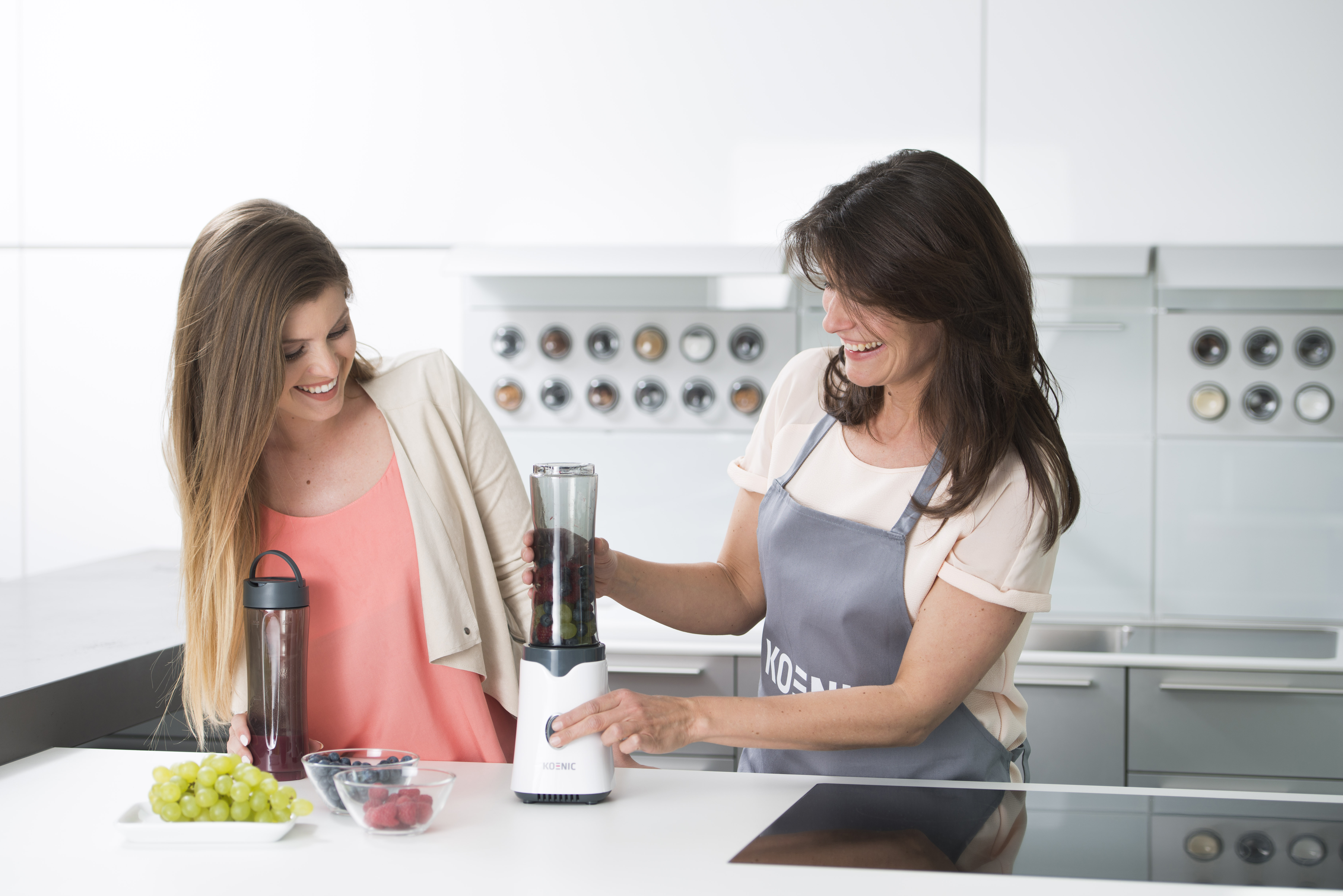 Zwei Frauen bereiten einen Smoothie zu. Eine Frau bedient einen Mixer, die andere lächelt und hält eine Flasche.