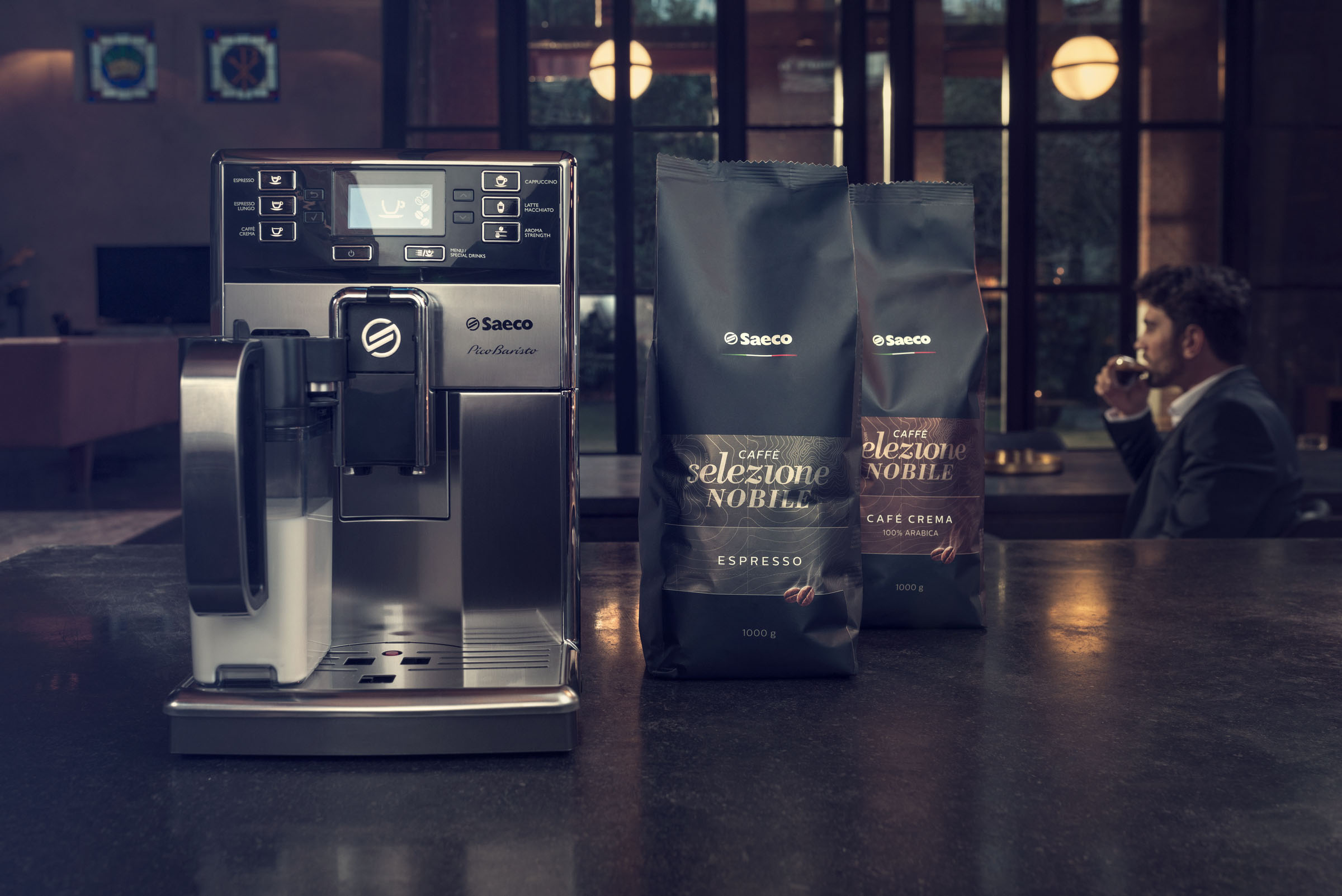 Kaffeemaschine, zwei Kaffeetüten und ein Mann, der in einem modernen, gedämpften Raum Kaffee trinkt.