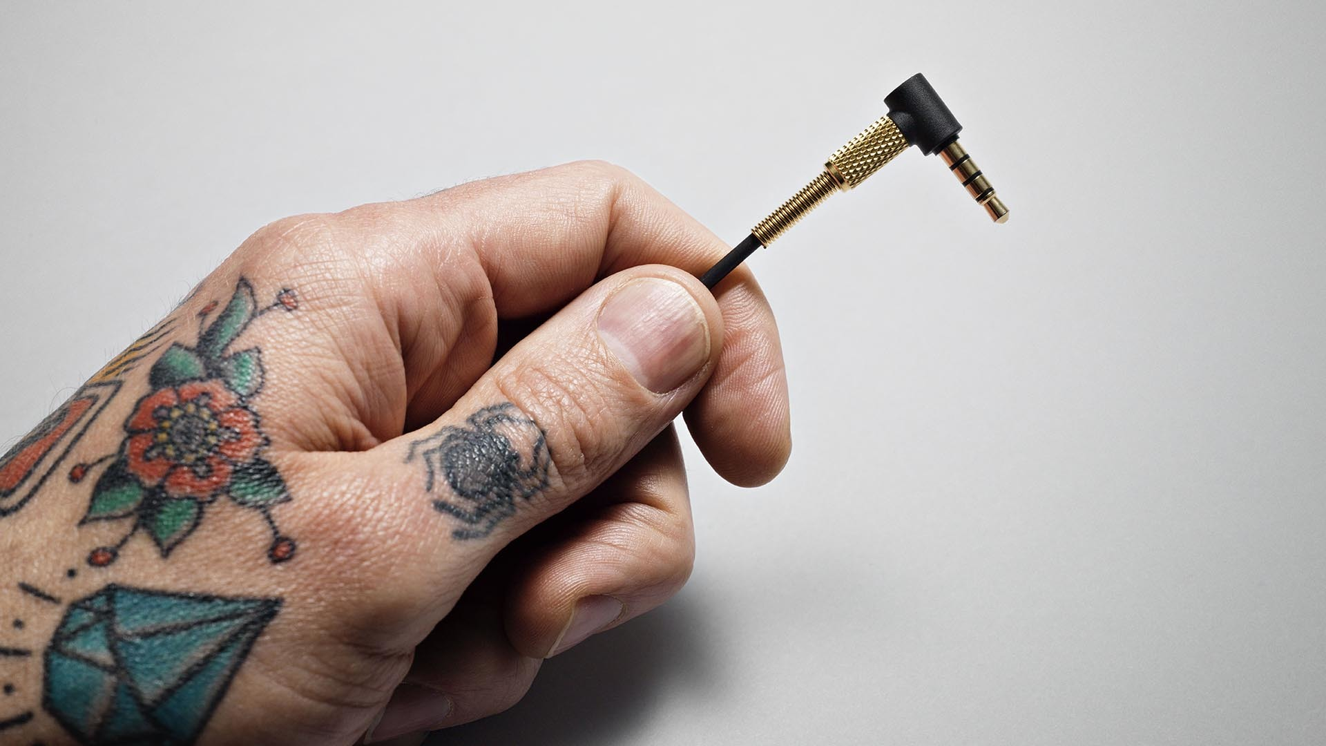Une main tatouée tient une prise audio 3,5 mm noire et dorée à pointe dorée.