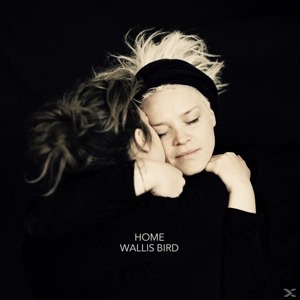 Zwei Frauen umarmen sich. Eine hat blonde Haare und ein schwarzes Stirnband. Text: 'HOME' und 'WALLIS BIRD'.