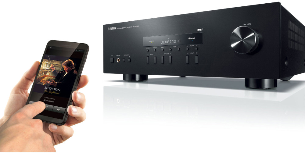 Schwarzer Stereo-Receiver mit einem schwarzen Smartphone, das Musikinformationen anzeigt. Hand berührt Telefon.