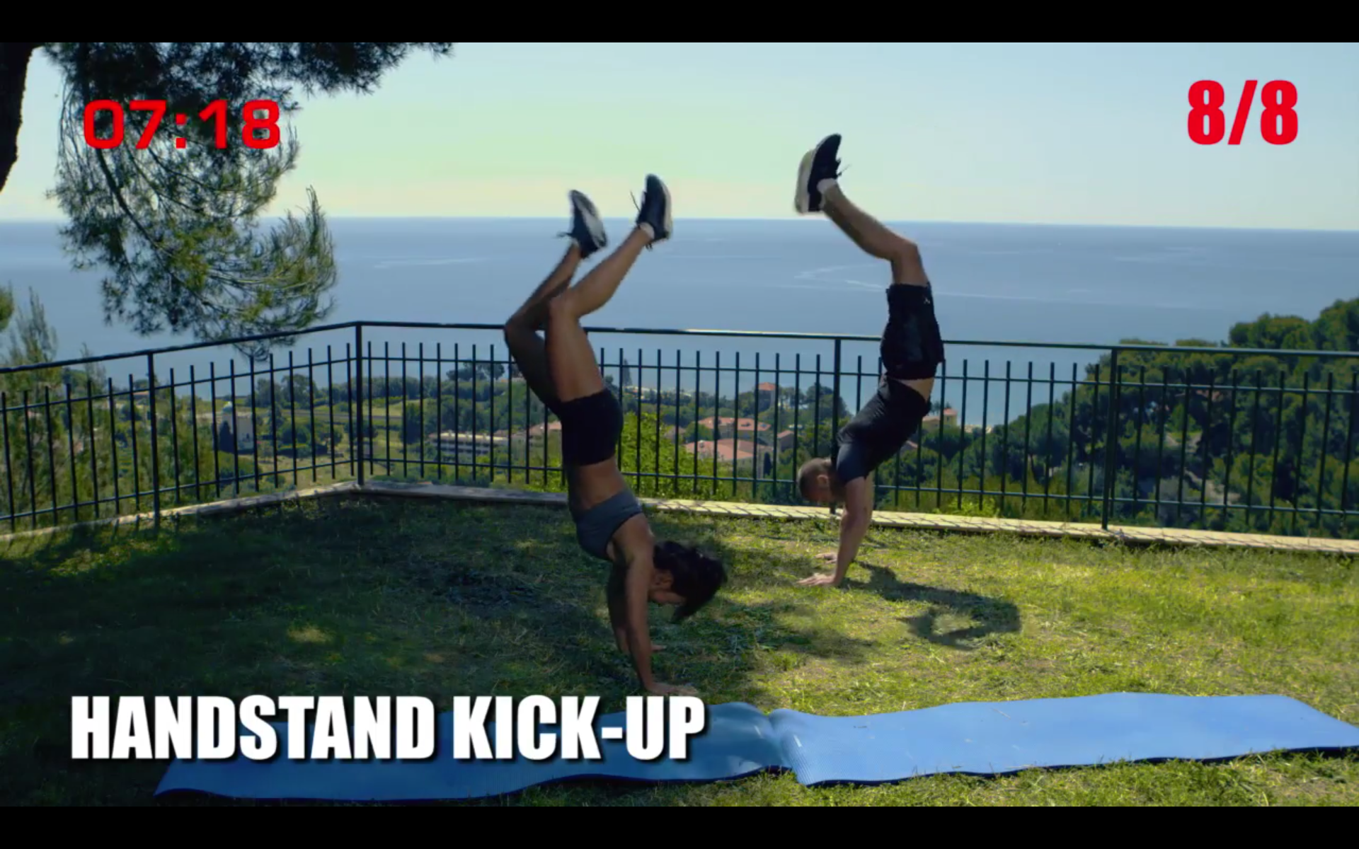 Zwei Personen machen draußen Handstand-Kick-Ups. Die Szene ist auf einem grasbewachsenen Hügel mit Blick auf das Wasser.