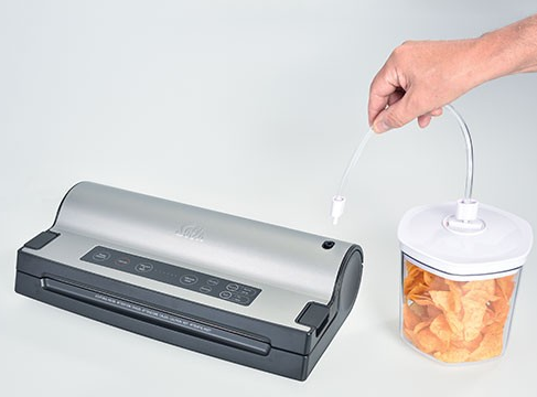 Een zilveren vacuüm sealer is verbonden met een bakje met chips. Een hand houdt de buis vast.