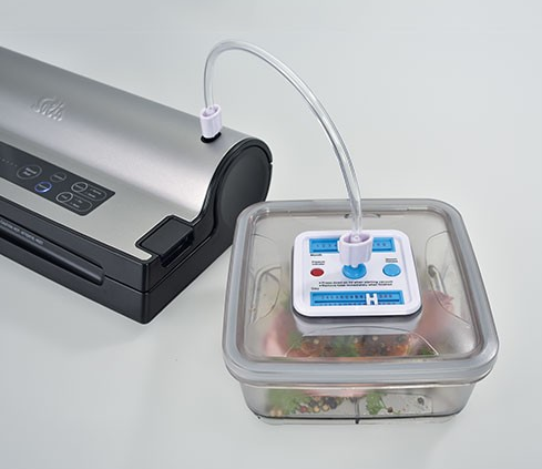 Vacuümsealer met bak verbonden door een buis. De bak bevat voedsel.