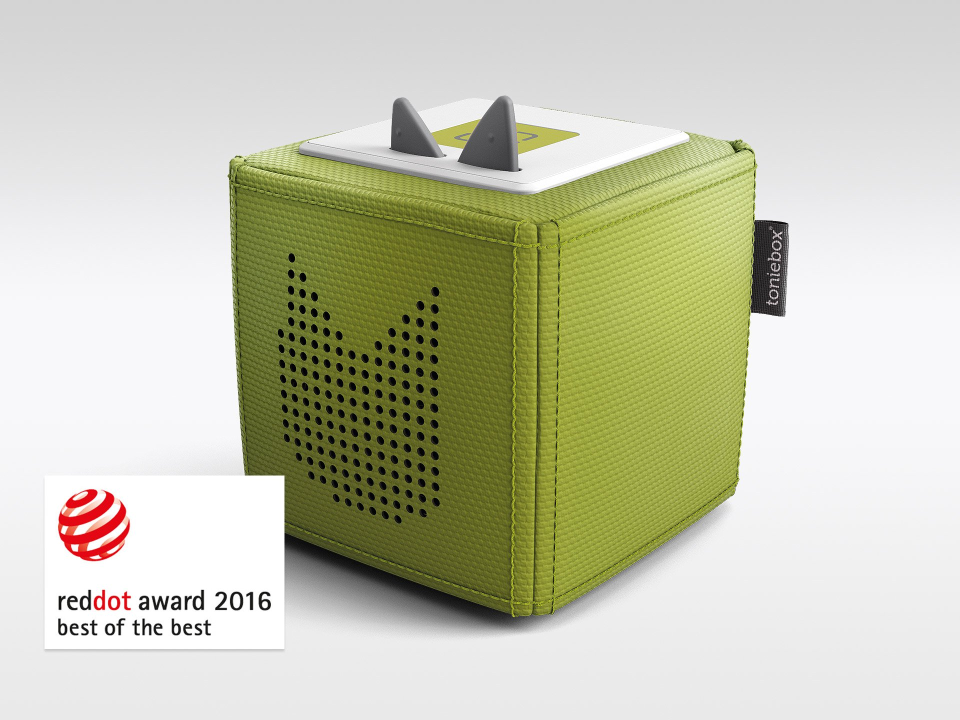 Grüner Würfel-Lautsprecher mit grauen Ohren, weißem Deckel und schwarzen Löchern. Red Dot Award Logo links unten.