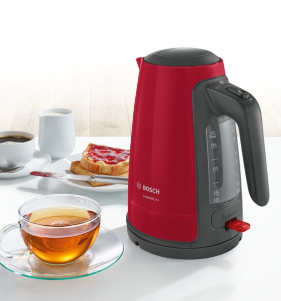 Roter Wasserkocher neben Tasse Tee, Toast mit Marmelade und weiteren Frühstückszutaten.