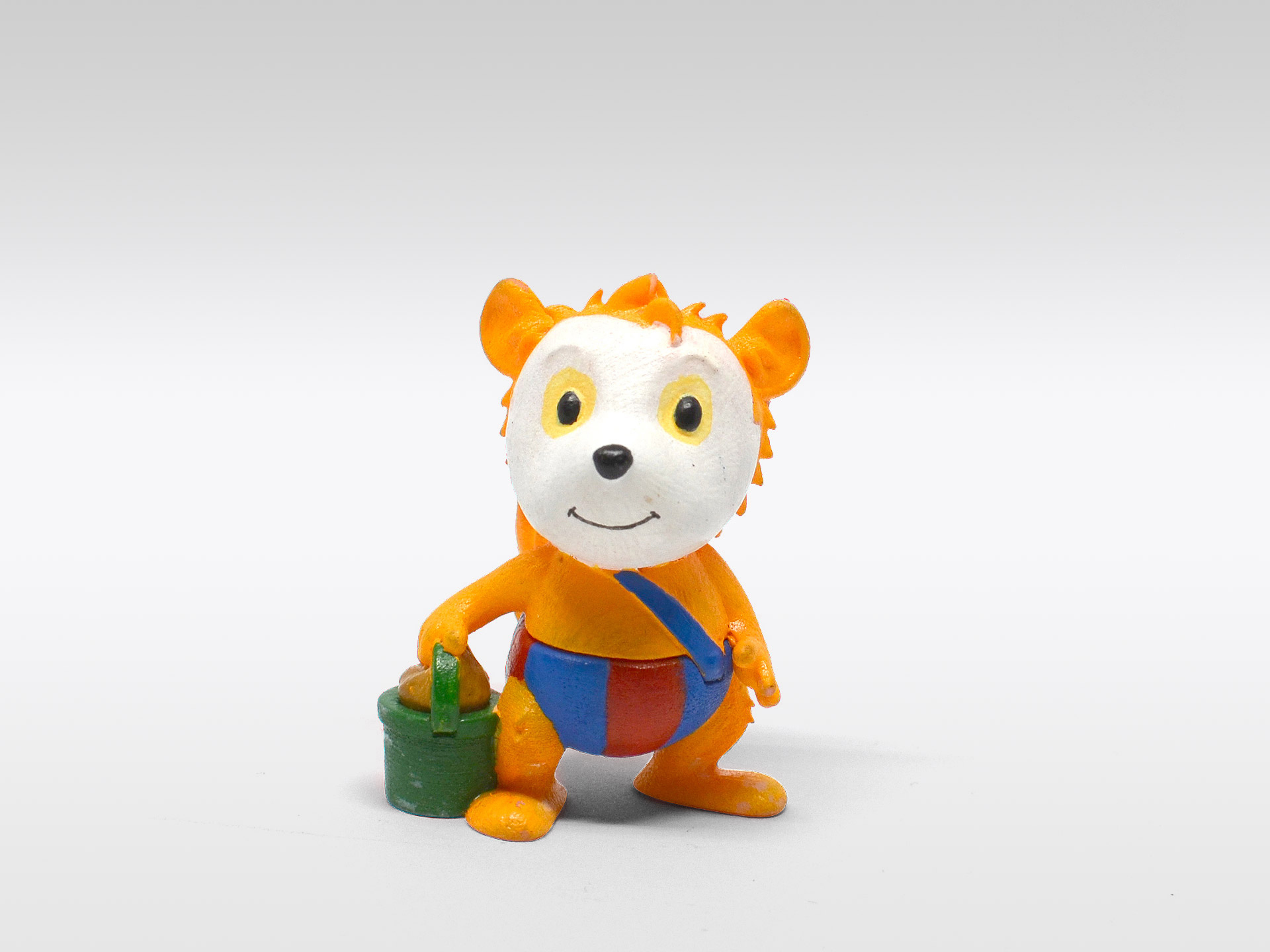 Eine kleine Bärenfigur mit orangefarbenem Haar, die einen grünen Eimer hält. Blaue und rote Shorts. Weißer Hintergrund.