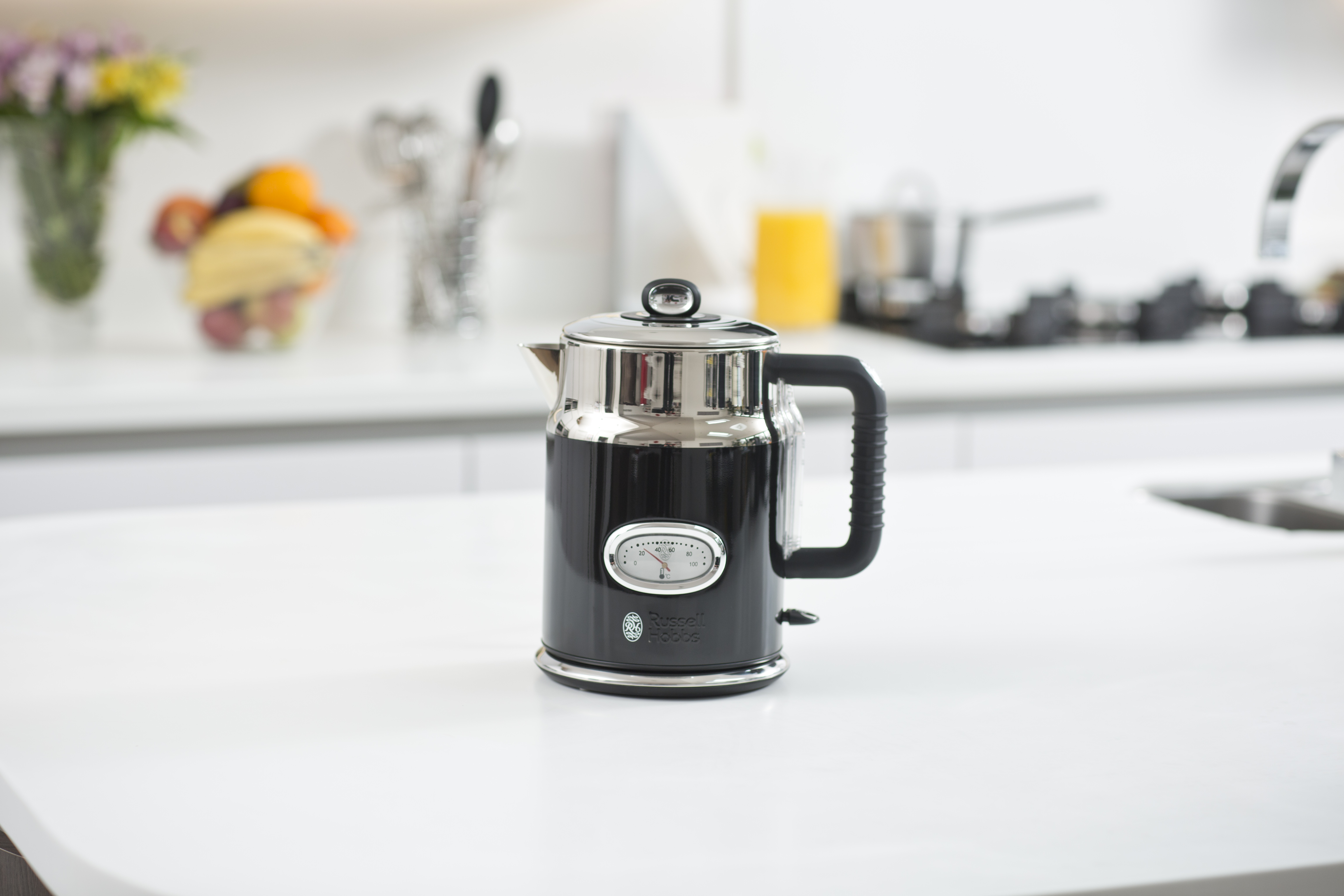 A black and silver kettle on a white counter, with flowers and a window in the background.