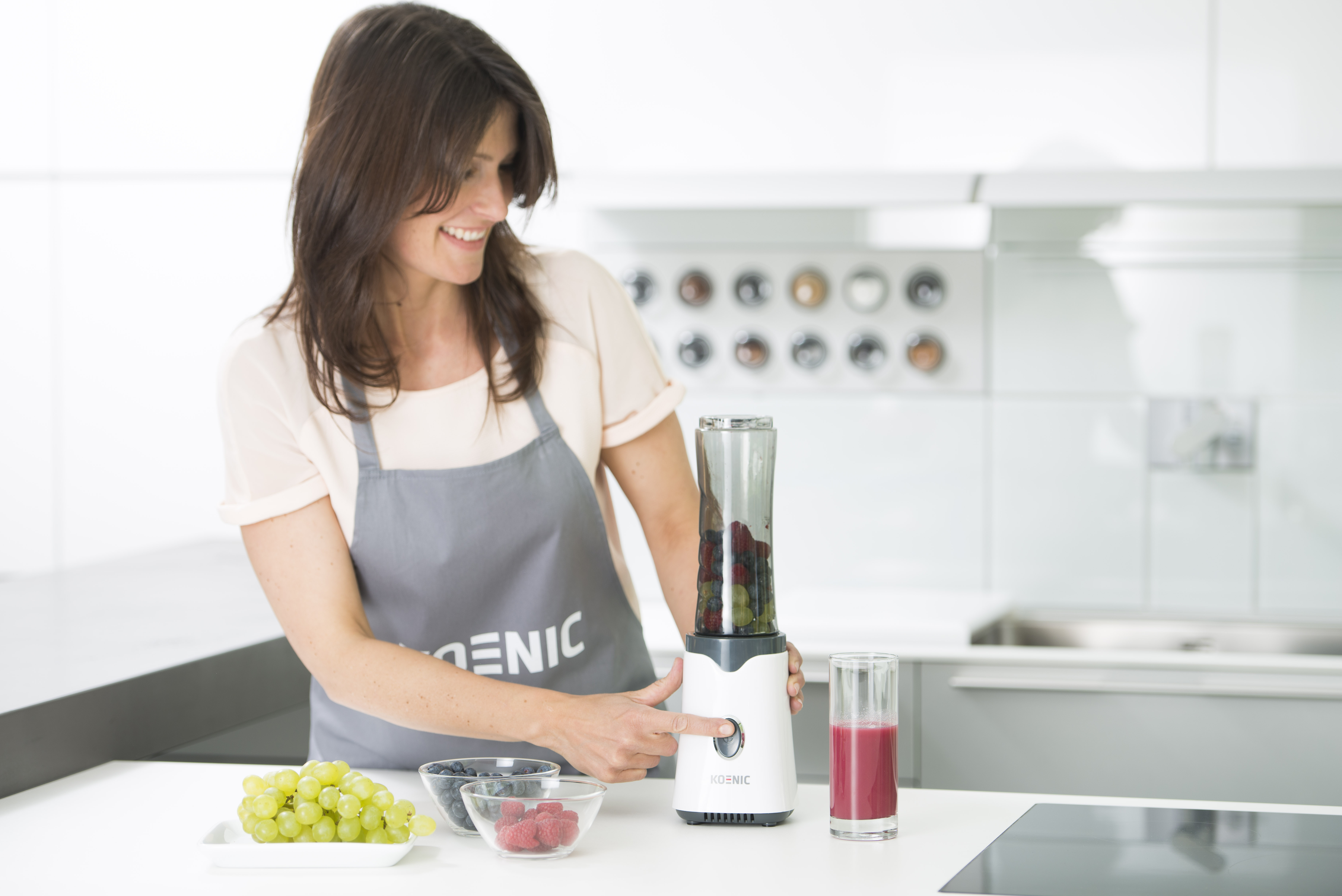 Frau in Küche, mixen Smoothie. Obst in Schalen, Mixer an, lächelnd.