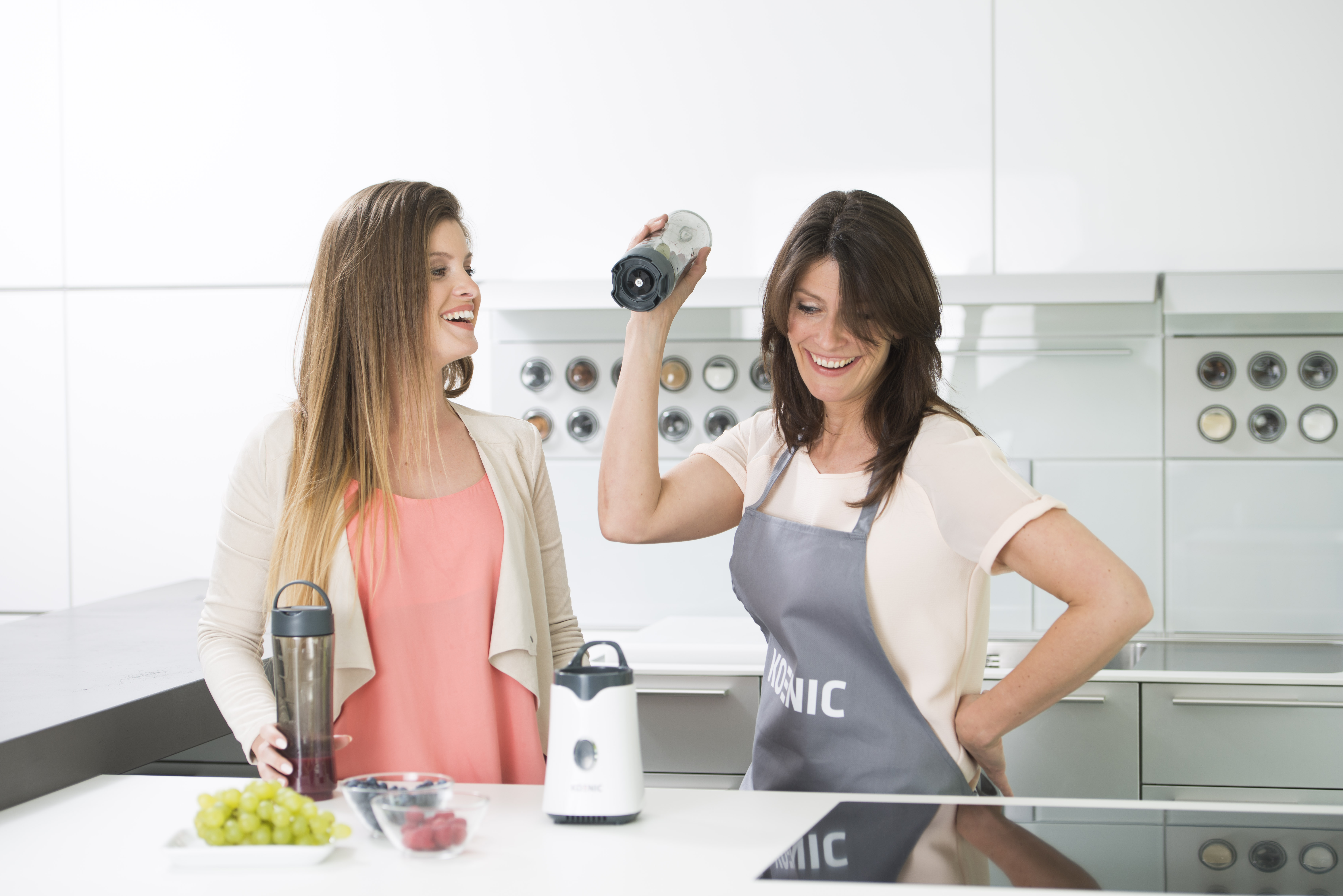 Zwei Frauen in einer modernen Küche, eine hält ein Mixerteil, lächelt.