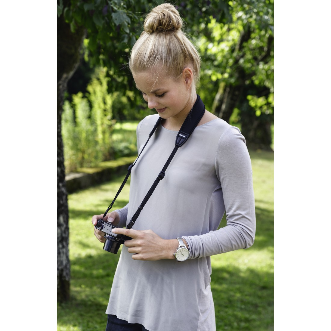 Femme blonde avec sangle d'appareil photo, tenant l'appareil, regardant vers le bas. Arbres verts en arrière-plan.