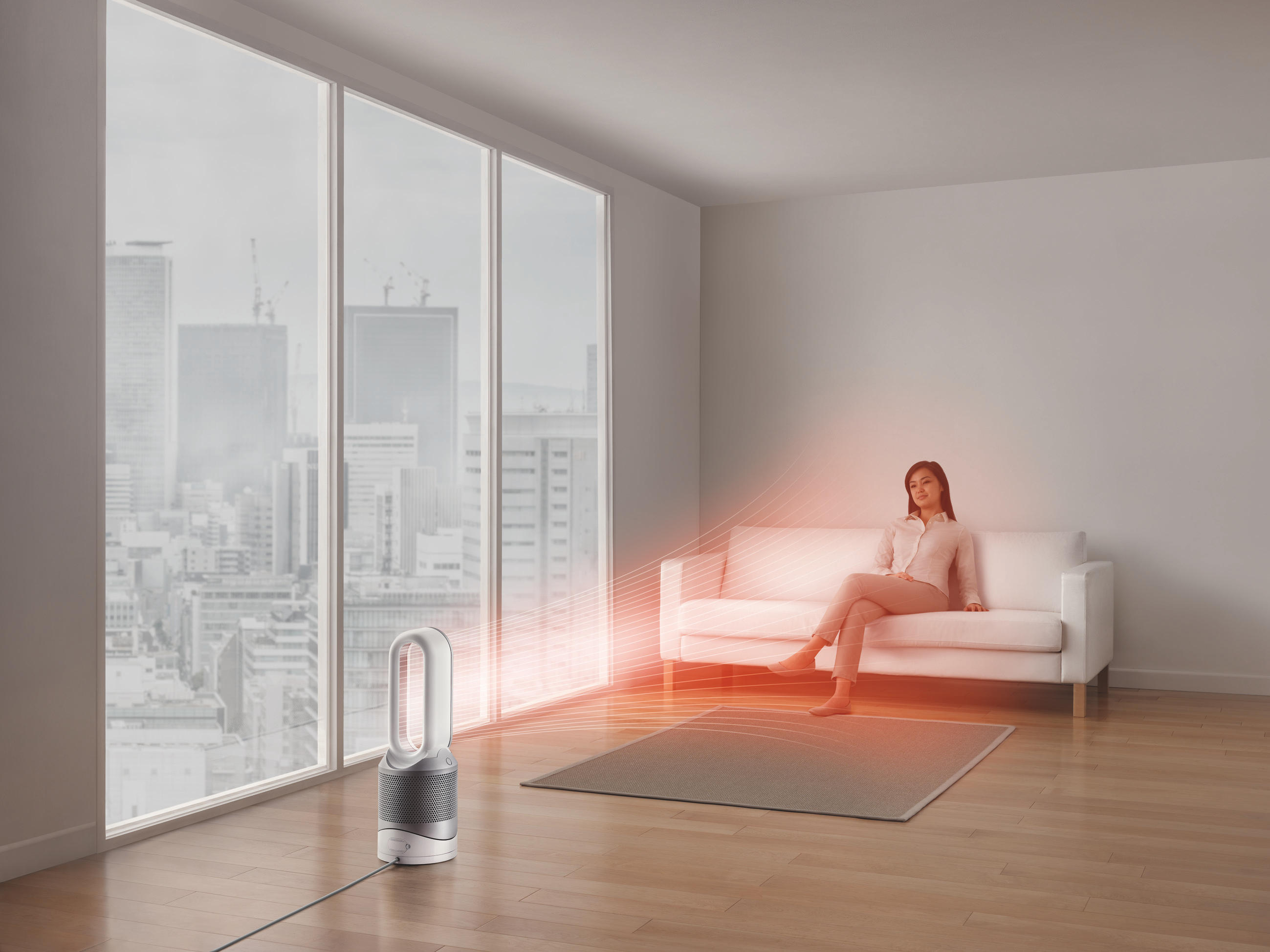 Eine Frau sitzt auf einem weißen Sofa in der Nähe eines großen Fensters, im Vordergrund ein Dyson-Ventilator.